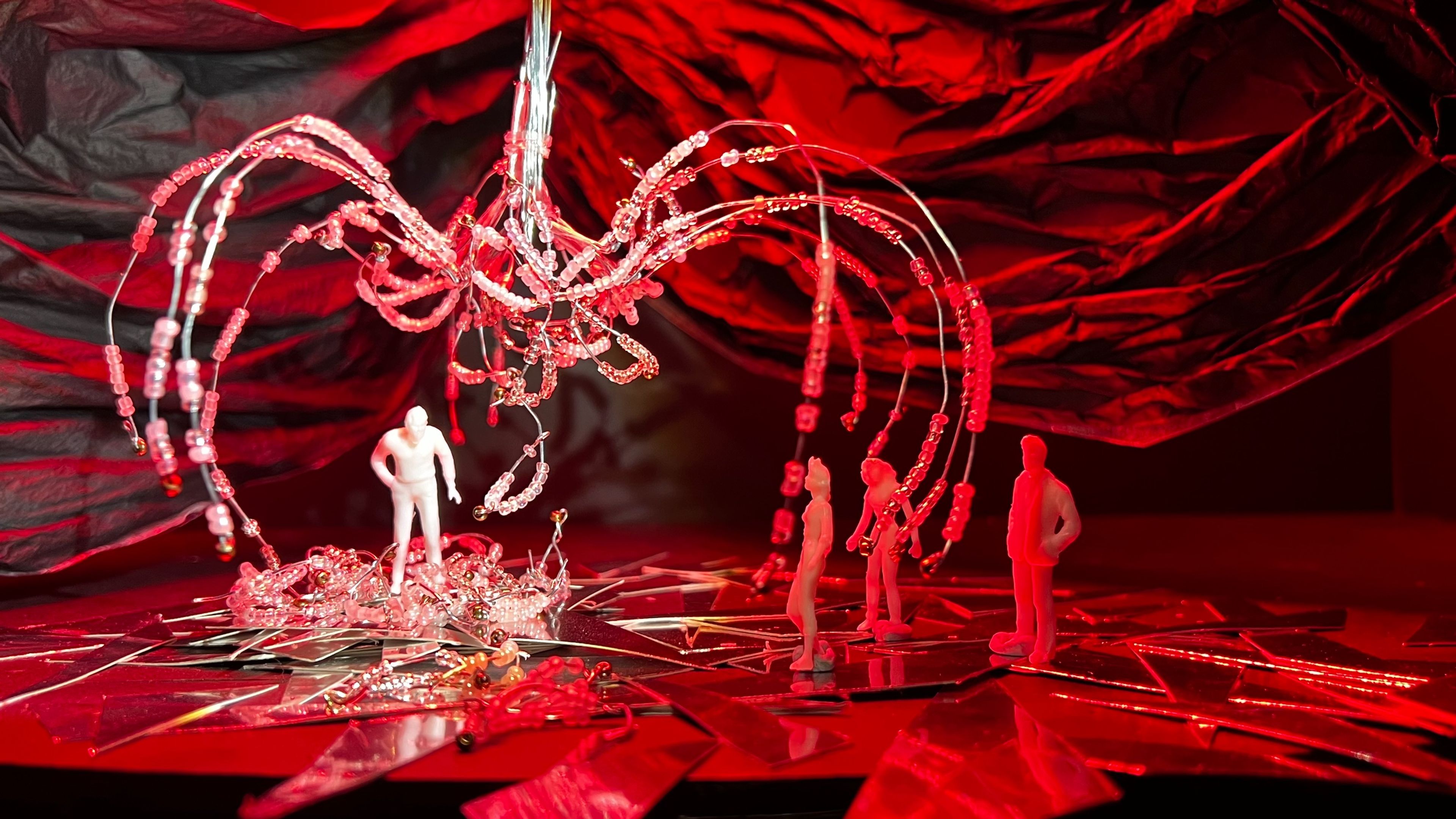 Theatre set model featuring white and red figures standing beneath a suspended beaded sculpture, with shattered mirror-like surfaces on the floor bathed in red light