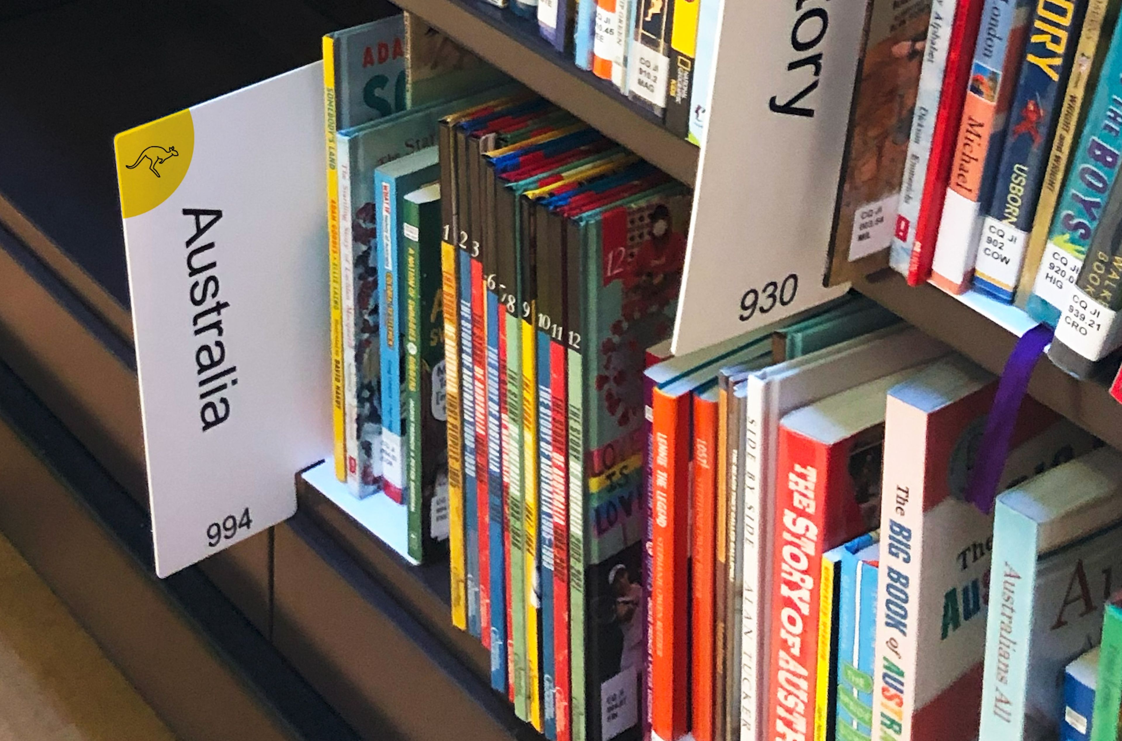 Library shelf with a white sign marked 'Australia' alongside a yellow circle featuring a kangaroo symbol. Books on the shelf are a mix of children's titles and Australian-themed publications, arranged in varying heights and colours, giving a lively, accessible look.