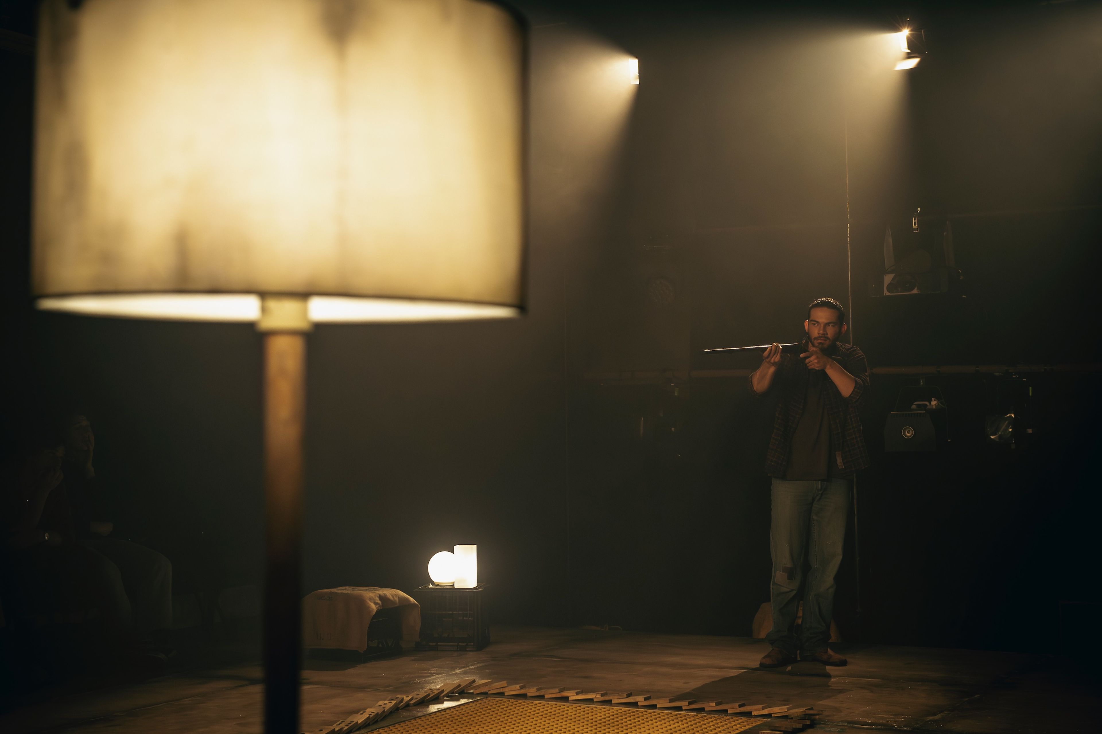 Production photograph of a dark theatre space with a performer aiming a prop rifle, lit by practical lamps creating pools of warm light