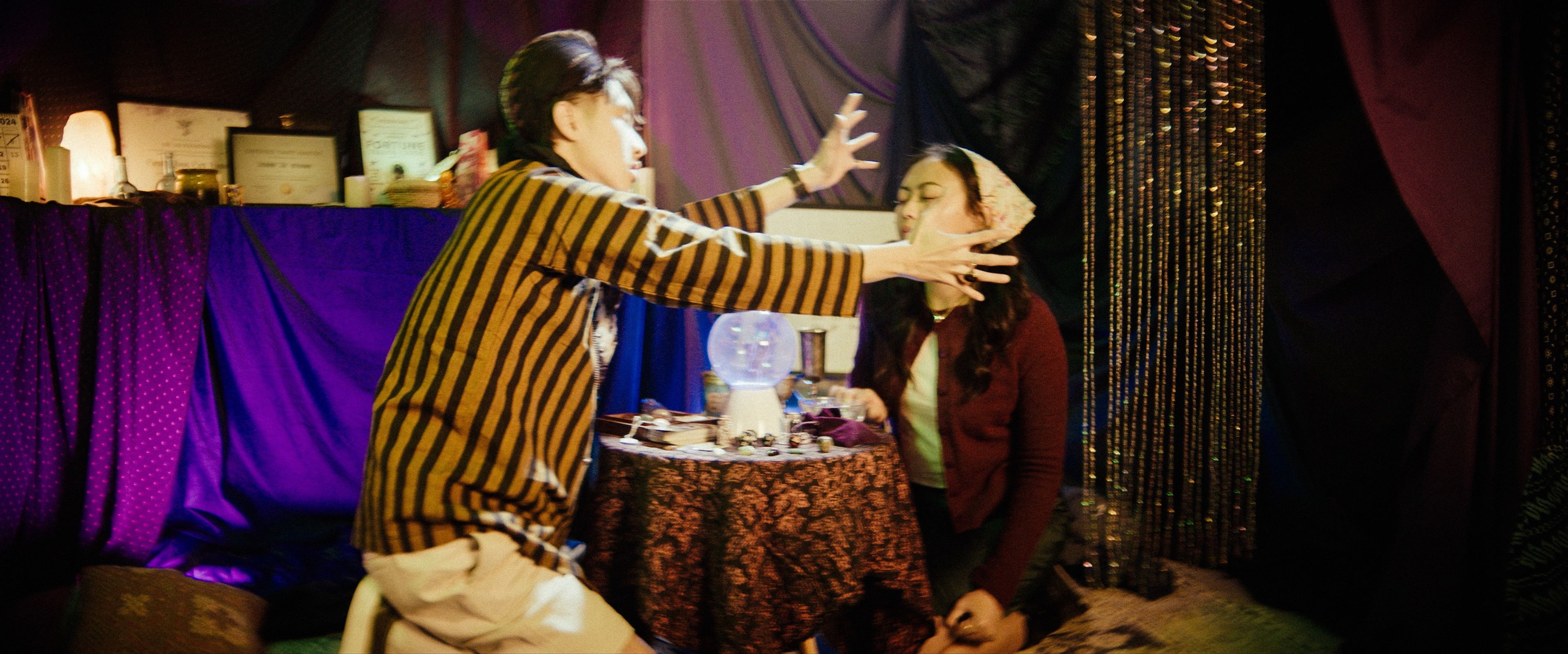 Film production still of a scene in a dimly lit fortune teller's room, with a performer in a striped robe making dramatic gestures towards another in maroon clothing, with mystical props and purple drapery in the background