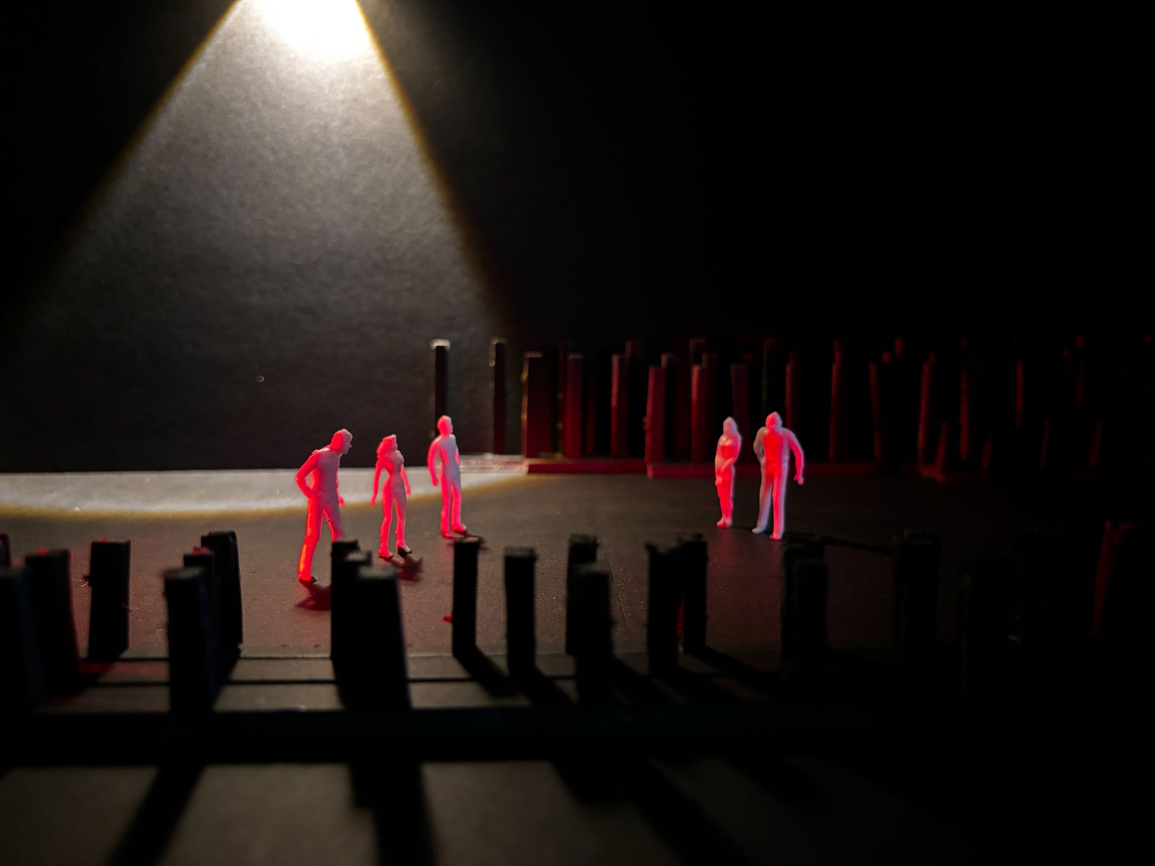 Stage model with five figures bathed in a warm spotlight, surrounded by vertical structures in a dark space.