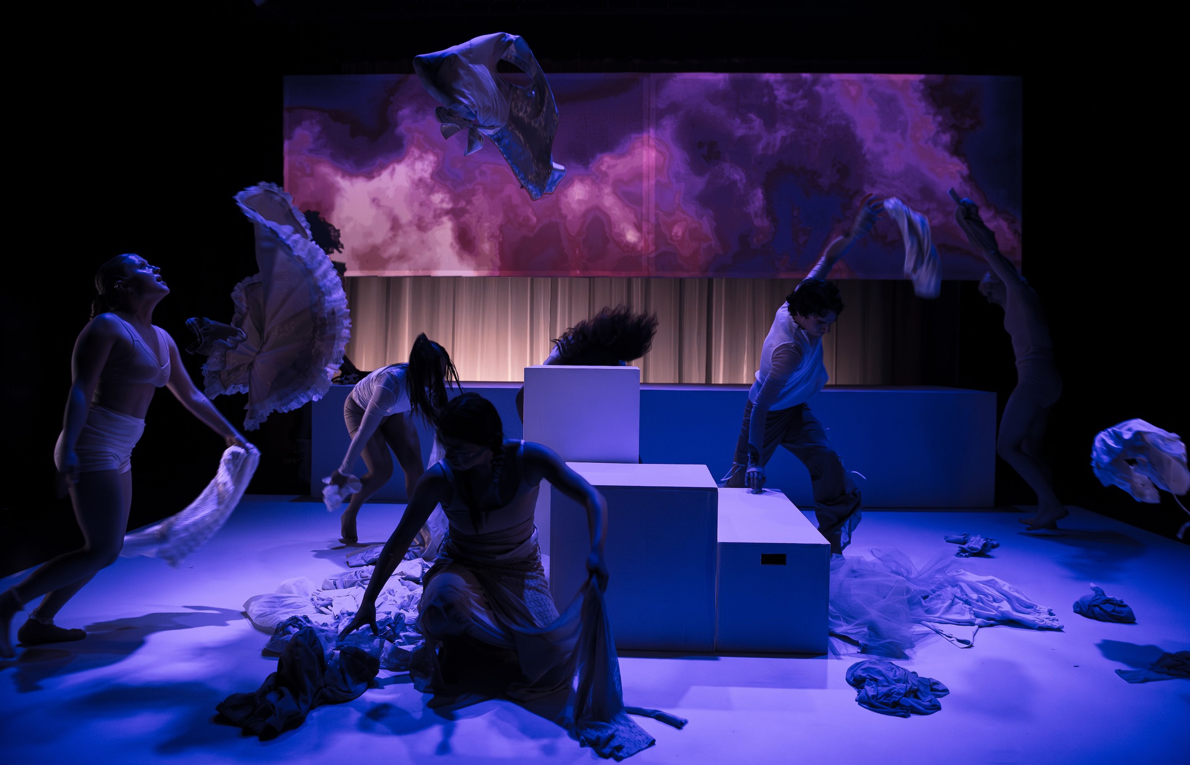 Theatre production photograph featuring a group of performers in white costumes, dancing dramatically with fabric flowing above, lit by purple and blue lighting.