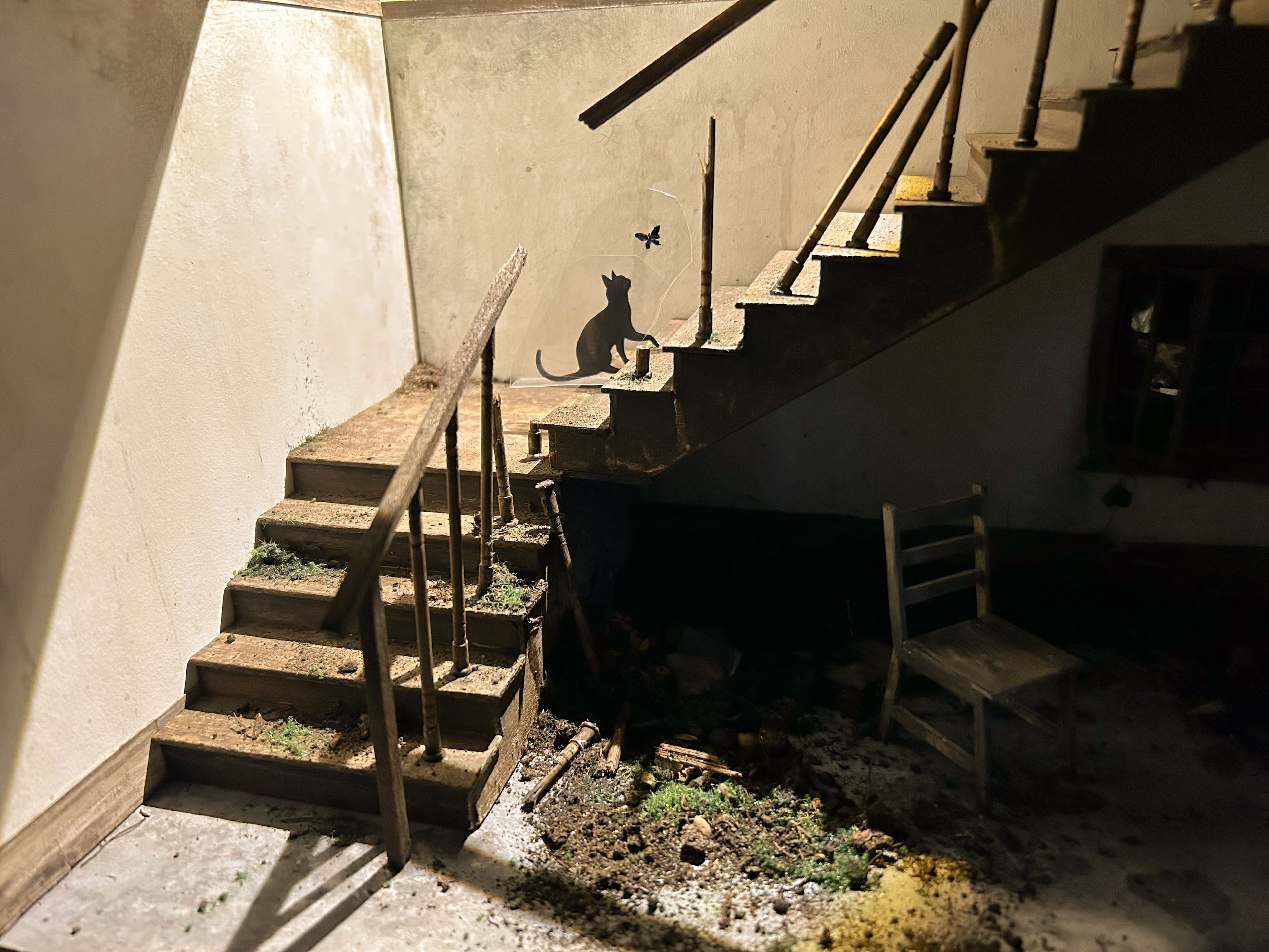 Diorama of a dimly lit, deteriorated stairwell with a rustic wooden chair at the bottom and a silhouette of a cat on the staircase. The worn textures and muted lighting give the scene an eerie, abandoned atmosphere.