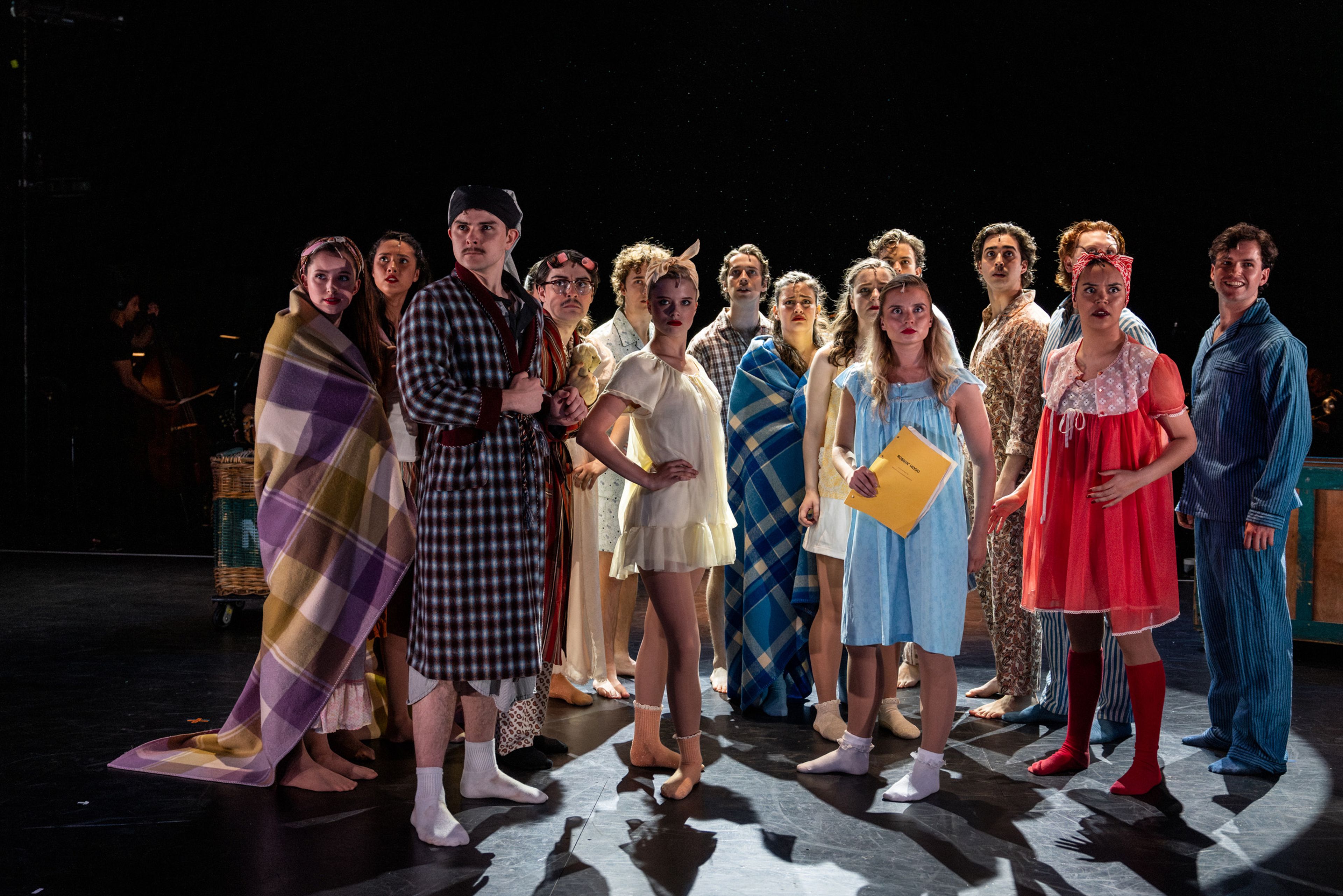 Theatre production photograph of performers in sleepwear and dressing gowns lined up on stage, with dramatic side lighting creating long shadows.