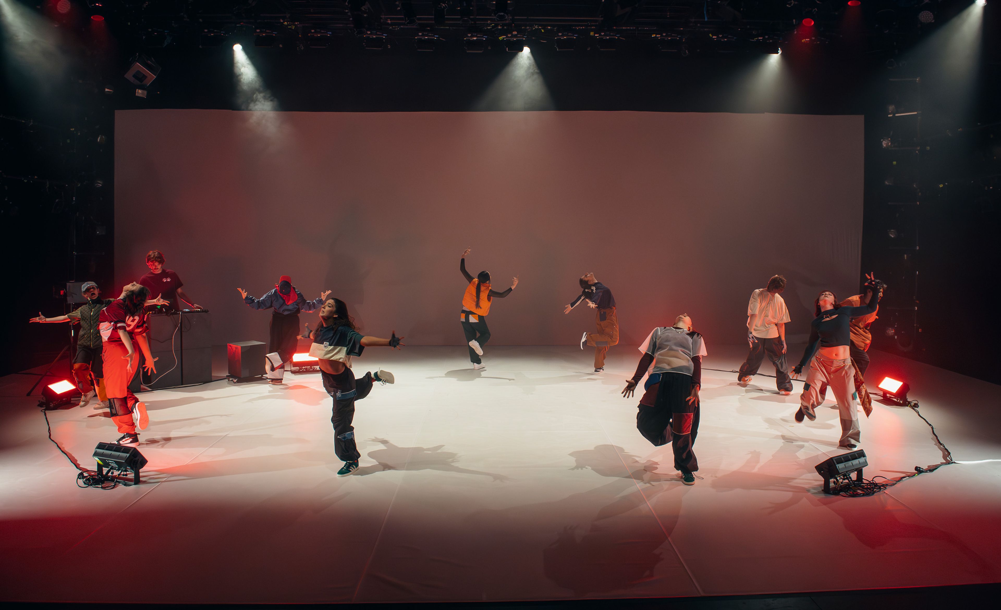 Dance production photograph of dancers spaced across the stage, each striking unique poses under spotlighting, with a DJ performing to the side.