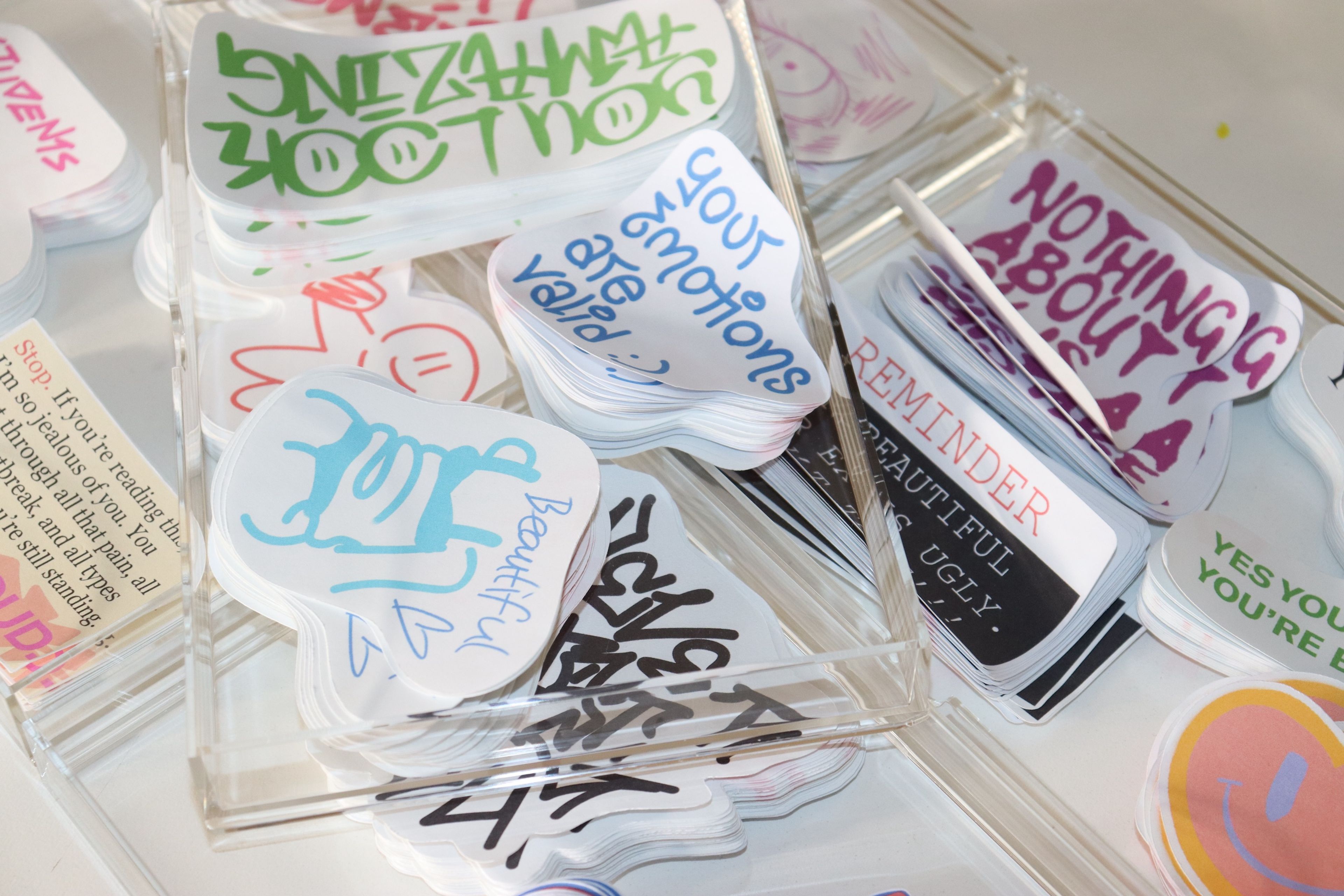 Collection of handmade stickers arranged in clear organisers, featuring encouraging messages with a mix of handwritten text and illustrations.