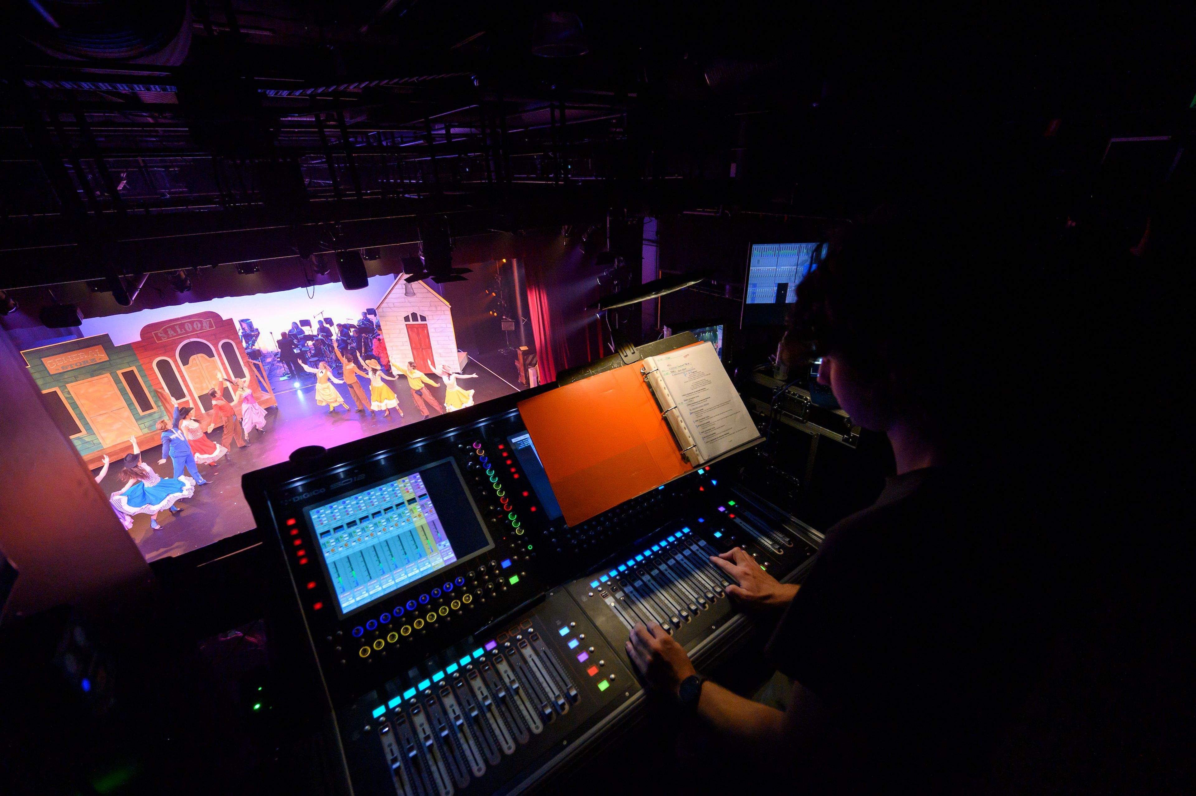 View from the control booth overlooking a stage with performers dancing, as a person operates the soundboard with a script open nearby.