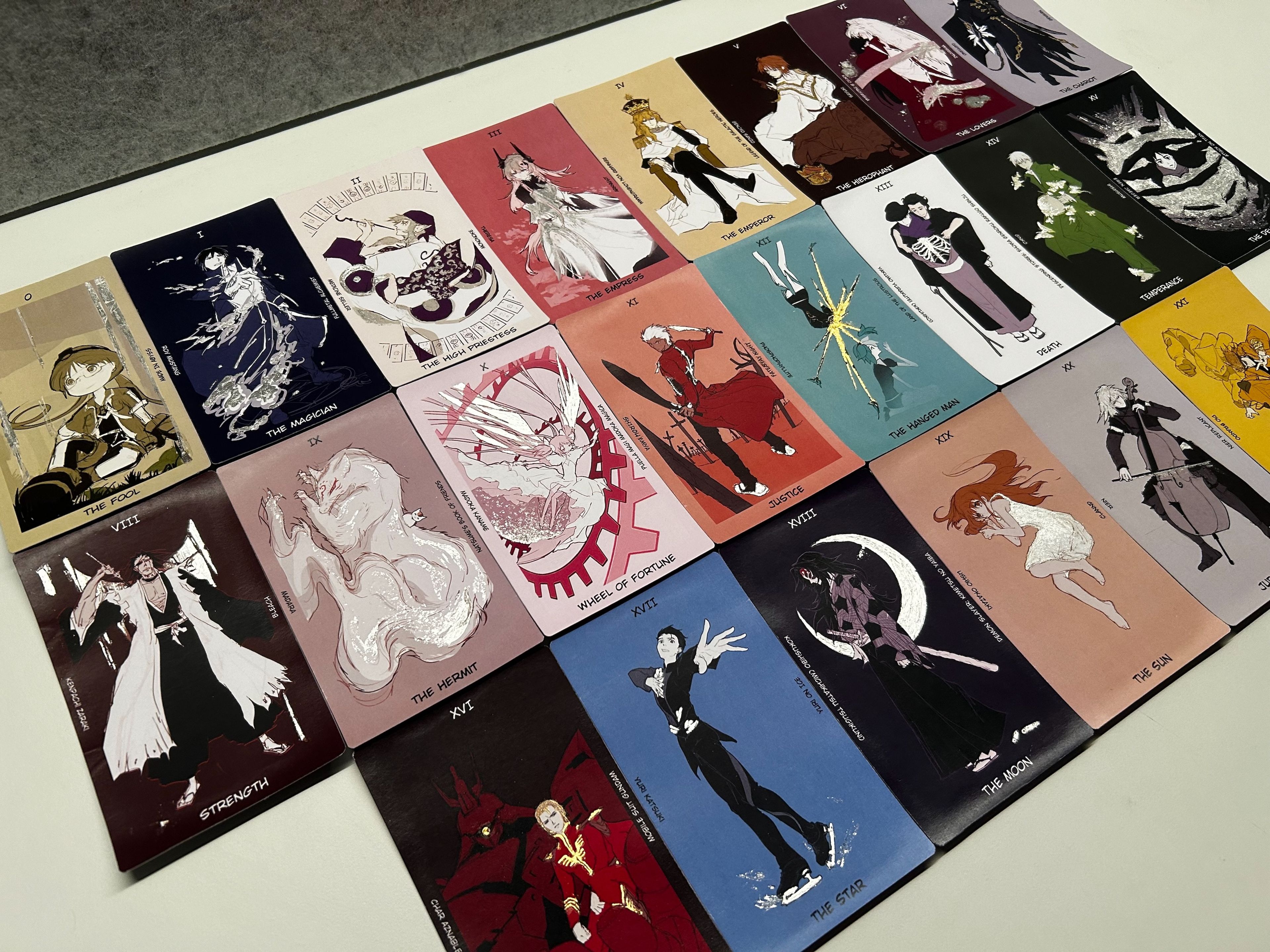 A collection of tarot-inspired cards arranged neatly in rows on a table, each card illustrated with unique characters and designs corresponding to tarot archetypes, such as "The Fool," "The Magician," and "The Lovers," each with distinct colour schemes and visual styles.