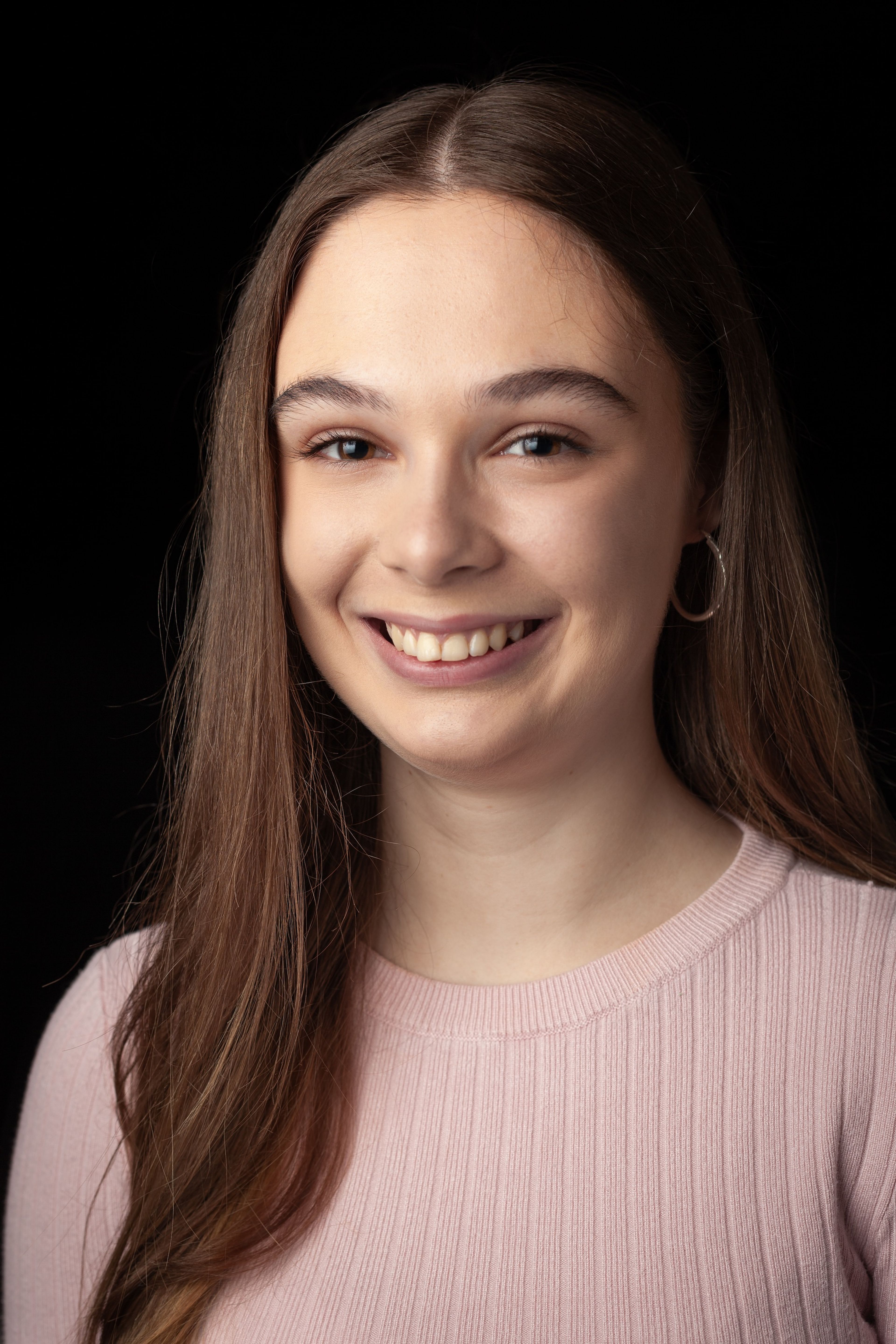 Headshot of a smiling young woman with long brown hair, wearing a light pink jumper and a hoop earring, against a black background.