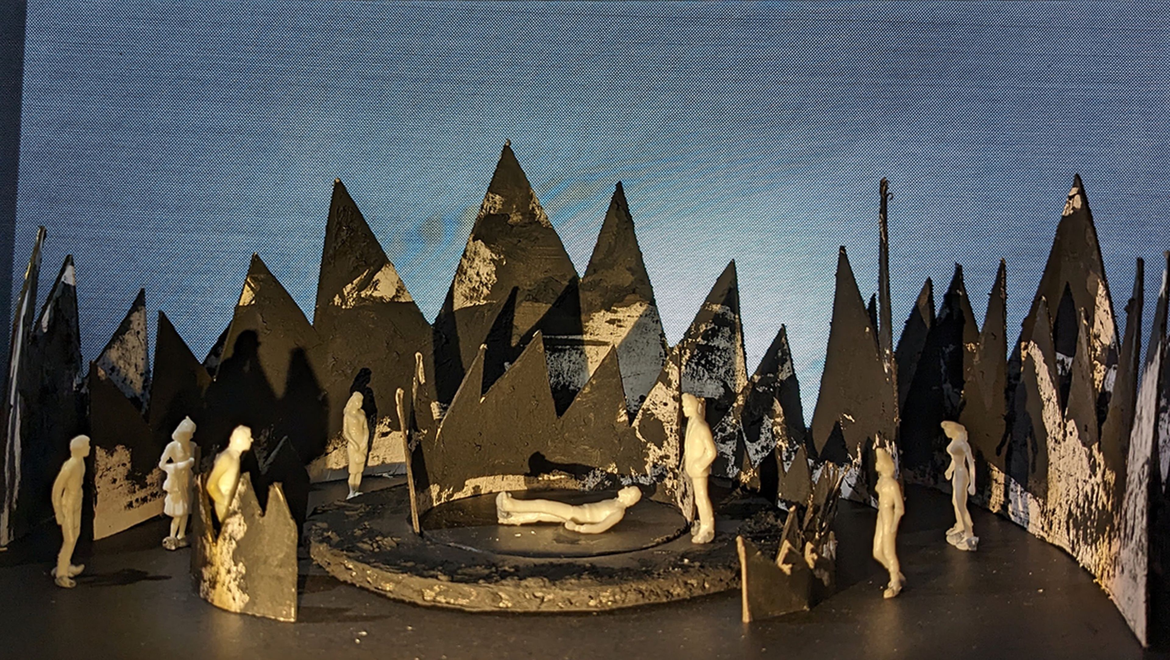 Set design model showing white figures interacting on a circular stage with a backdrop of black, jagged mountains and a blue gradient sky.