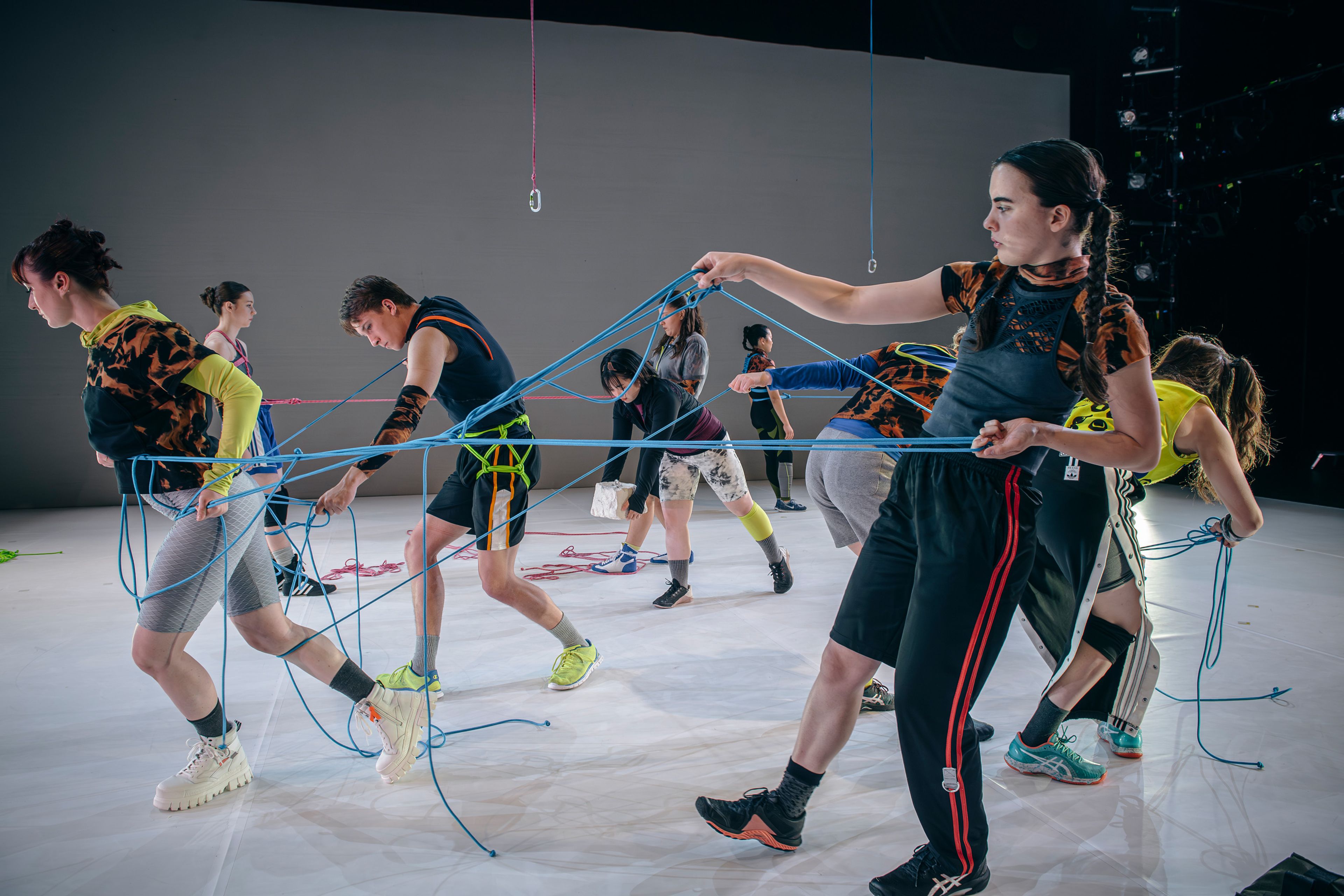 Dance production photograph of multiple dancers working with ropes, creating a web-like structure, with each performer pulling on different ropes in choreographed tension.