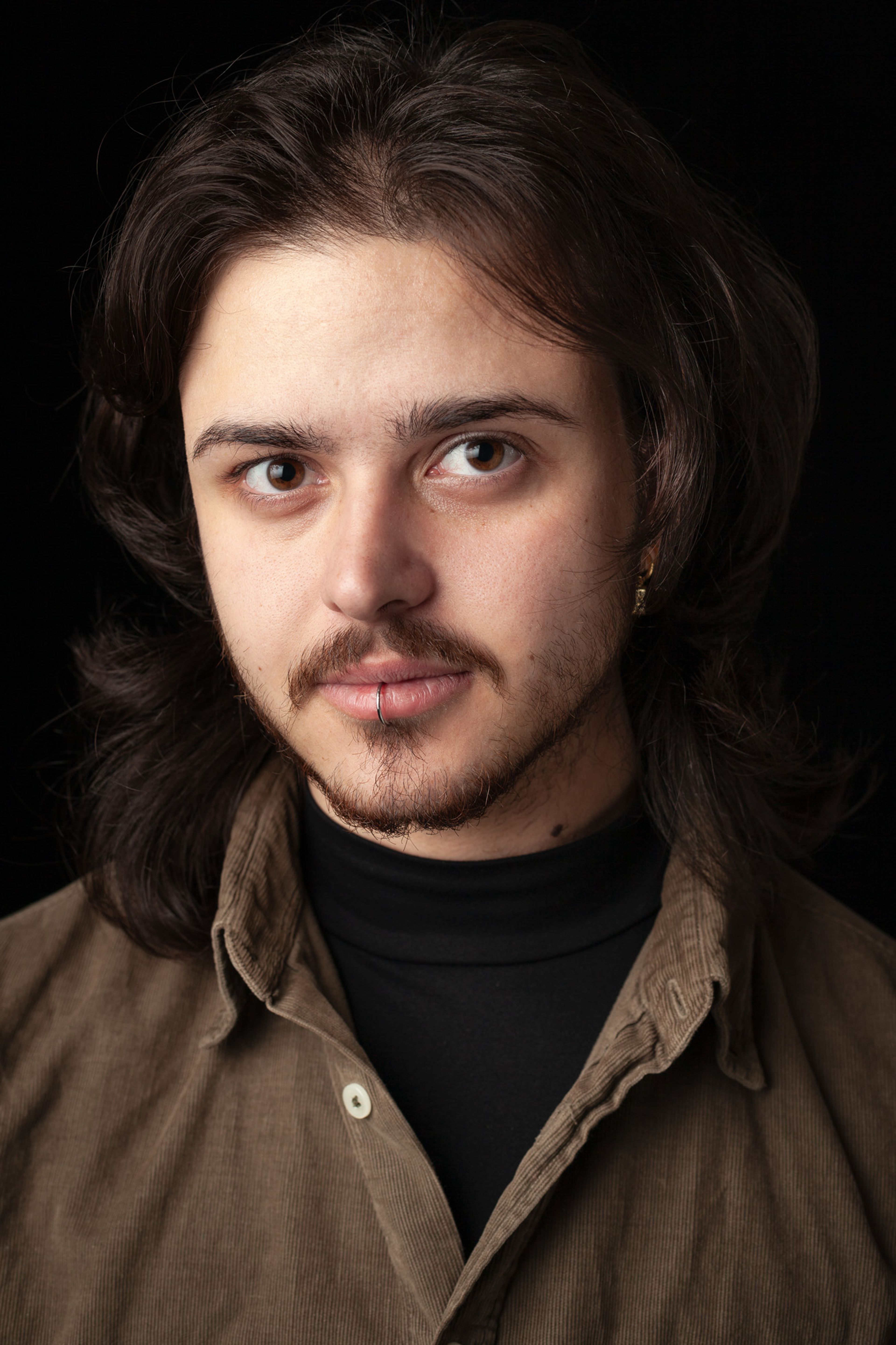 Headshot of Oliver who has with shoulder-length dark hair, a moustache, and a lip ring. They are wearing a brown button-up shirt, against a black background.