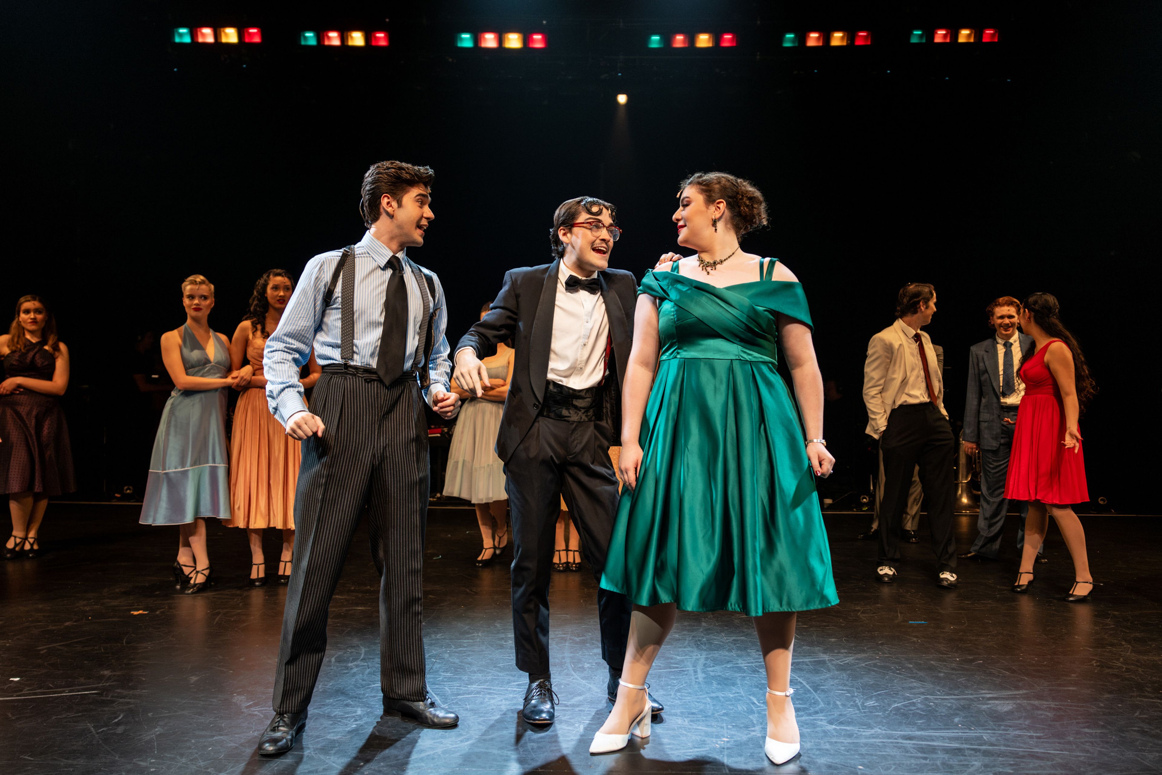 Theatre production photograph of three performers in 1950s formal wear - two men in suits and a woman in an emerald green dress - sharing a moment of laughter while other cast members are visible in the background.