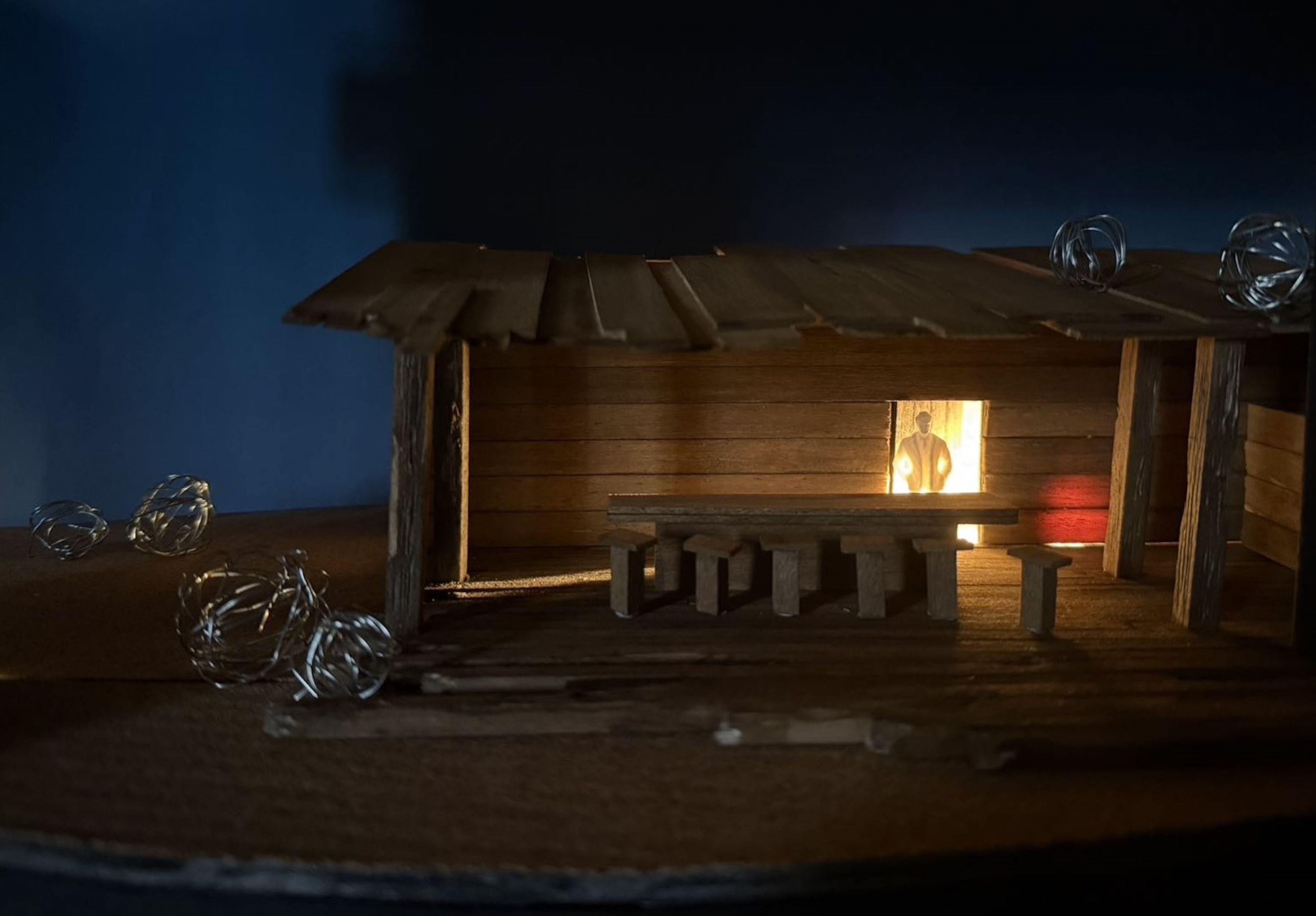 Scale model of a wooden saloon set design, with a lit figure visible through a window, set in a darkened space with metal wire tumbleweeds on the ground.