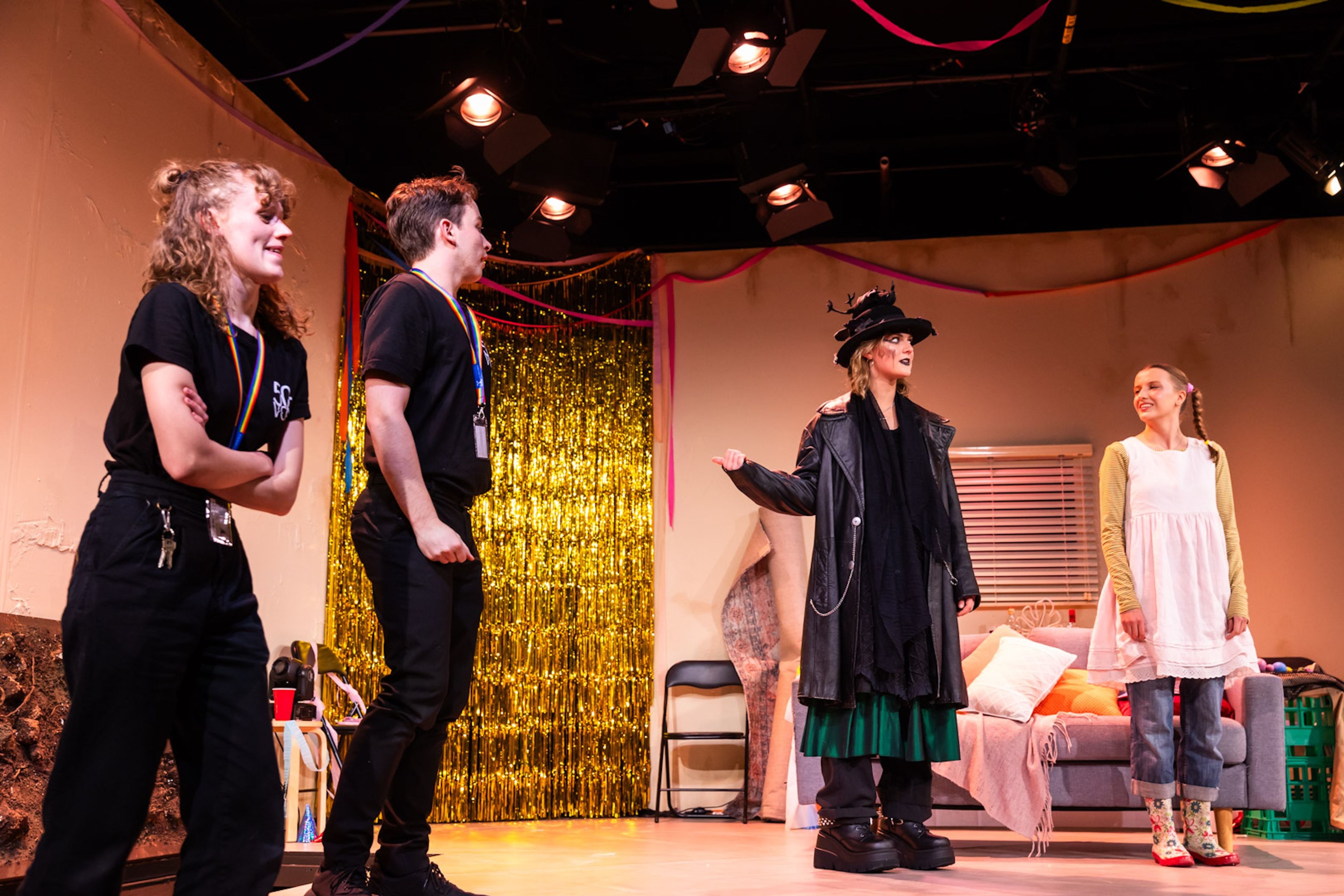 Theatre production photograph showing stage crew in black uniforms observing performers, one in a dramatic black coat and hat, against a backdrop with gold tinsel curtain and living room set.