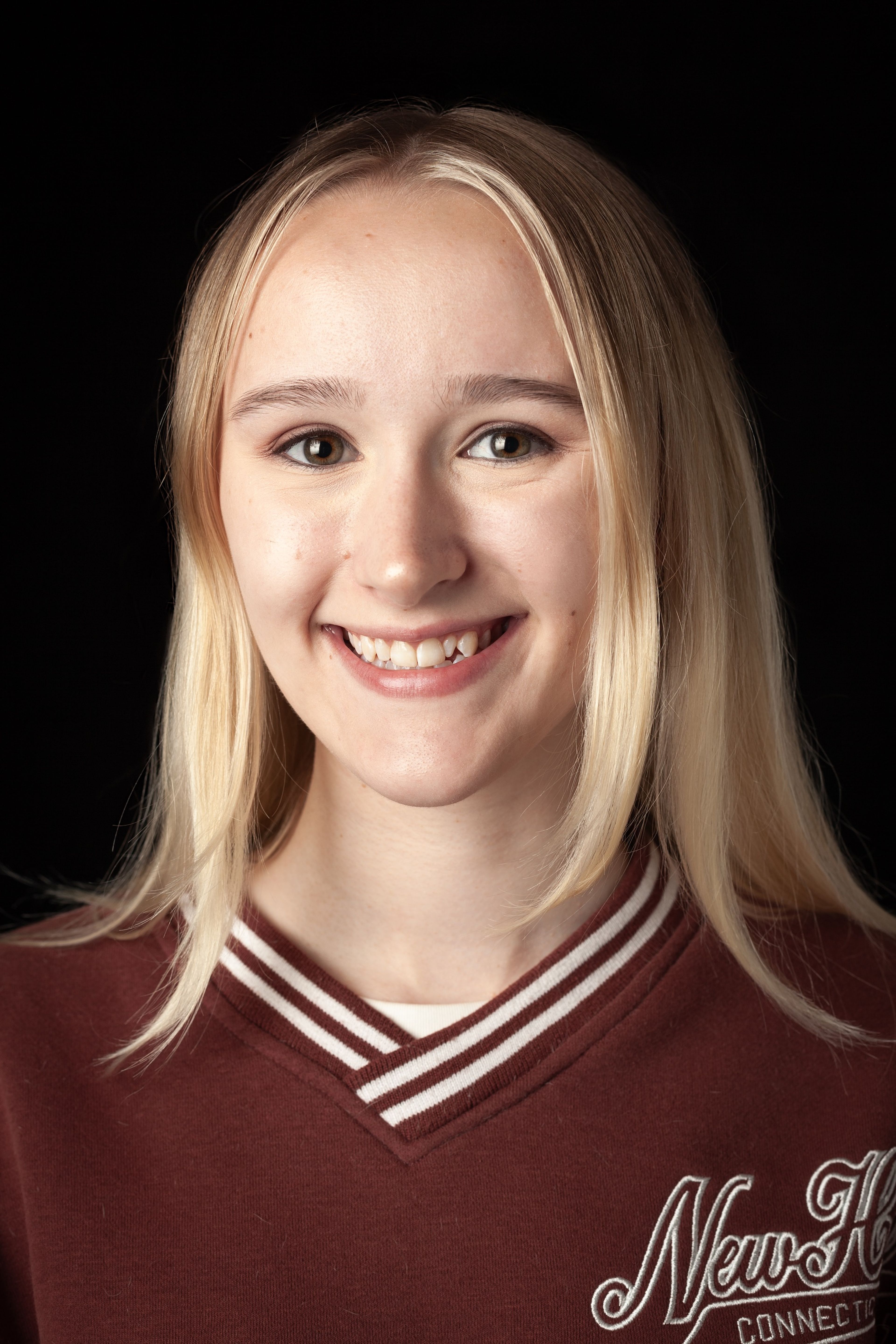 Headshot of a smiling young woman with straight blonde hair, wearing a maroon sweatshirt, against a black background.