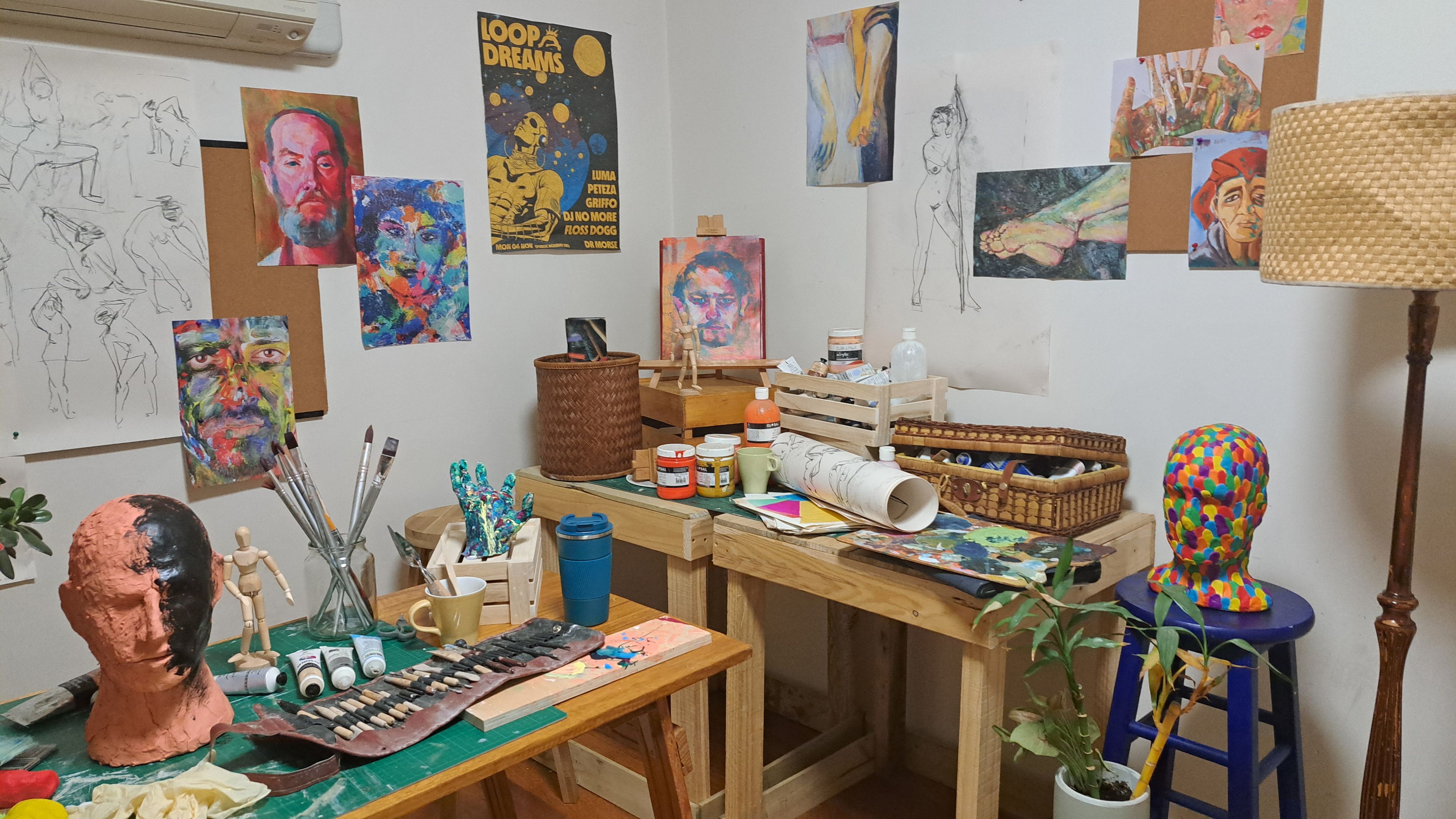 Set dressing photograph showing an artist's workspace with colourful paintings and sketches on the walls, various art supplies on the tables, including paint jars, brushes, a sculpted head, and a multicoloured mannequin head on a stool.