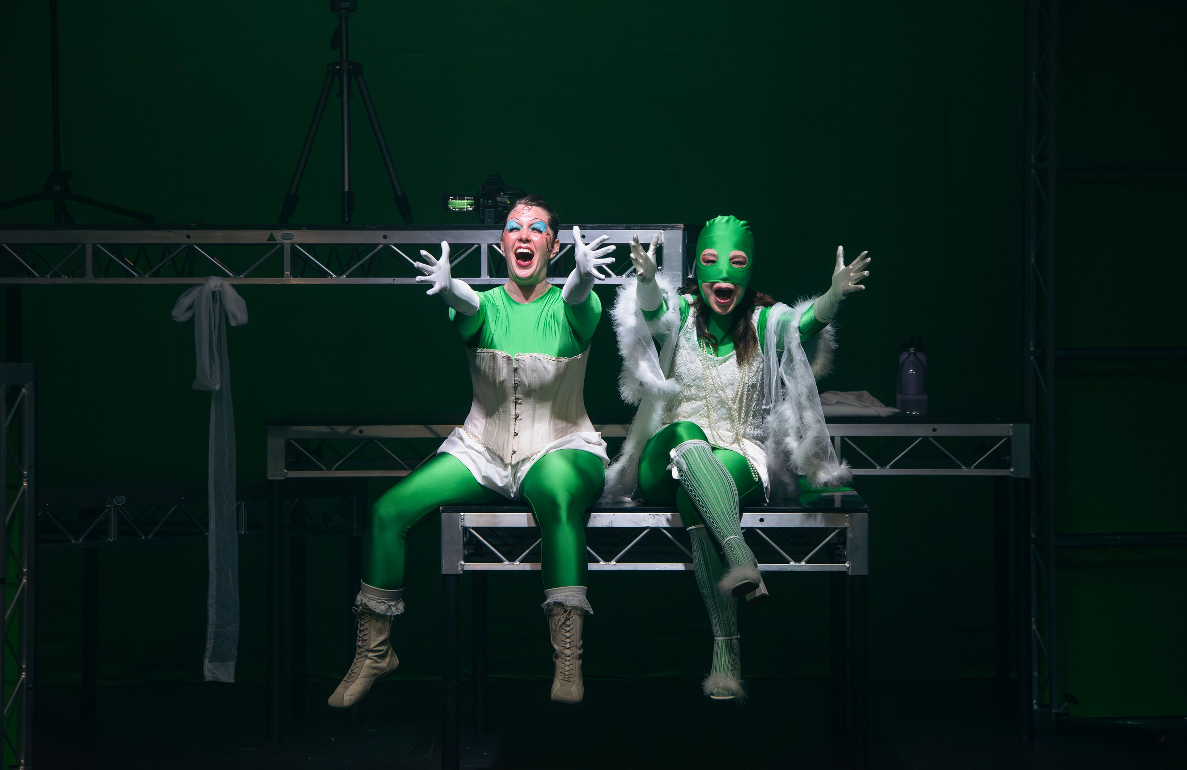 Theatre production photograph showing two actors dressed in bright green costumes, seated side by side on stage with animated expressions and outstretched arms, set against a dark green background.