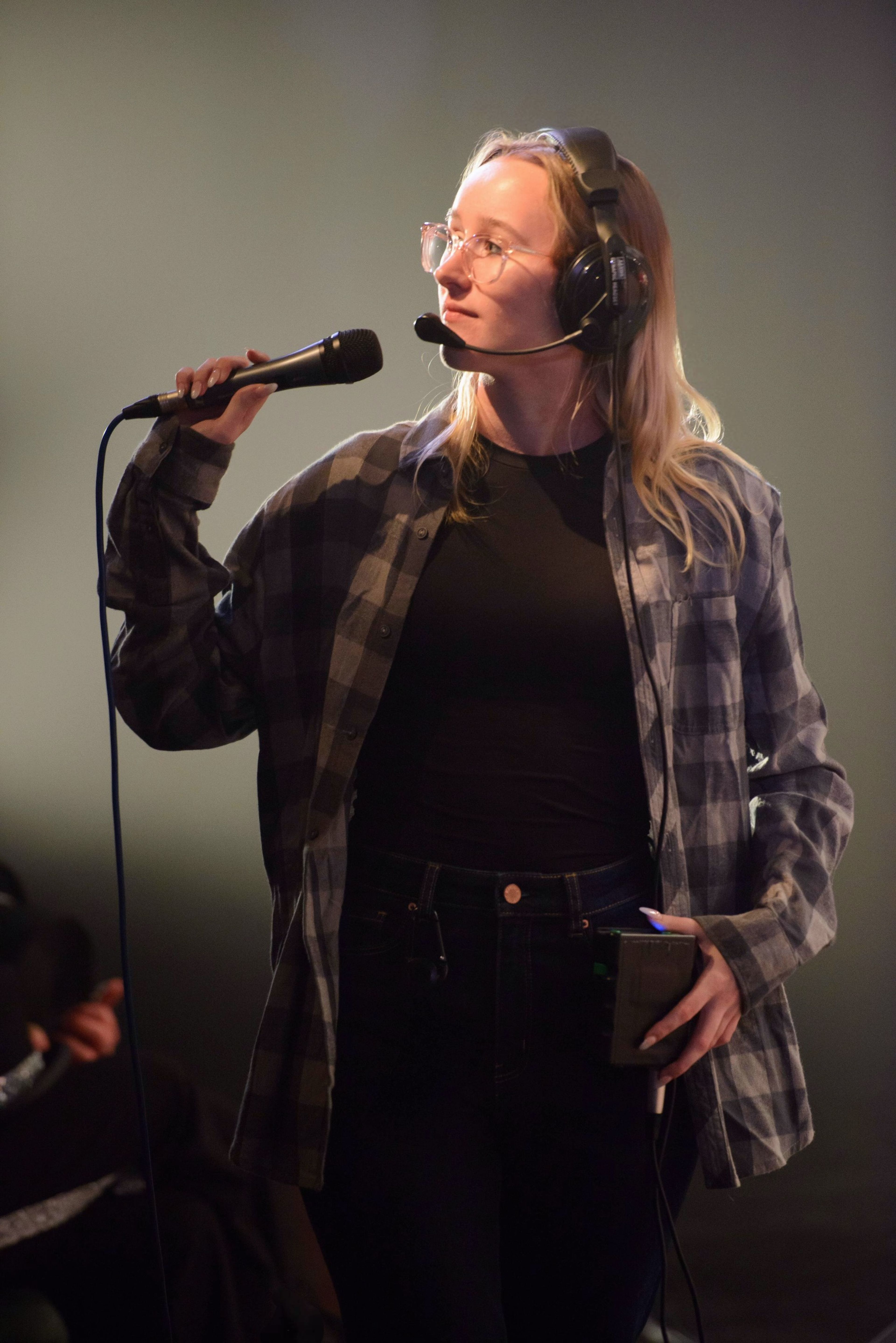 A woman in a plaid shirt and black t-shirt wearing a headset and holding a microphone, running a technical rehearsal onstage.