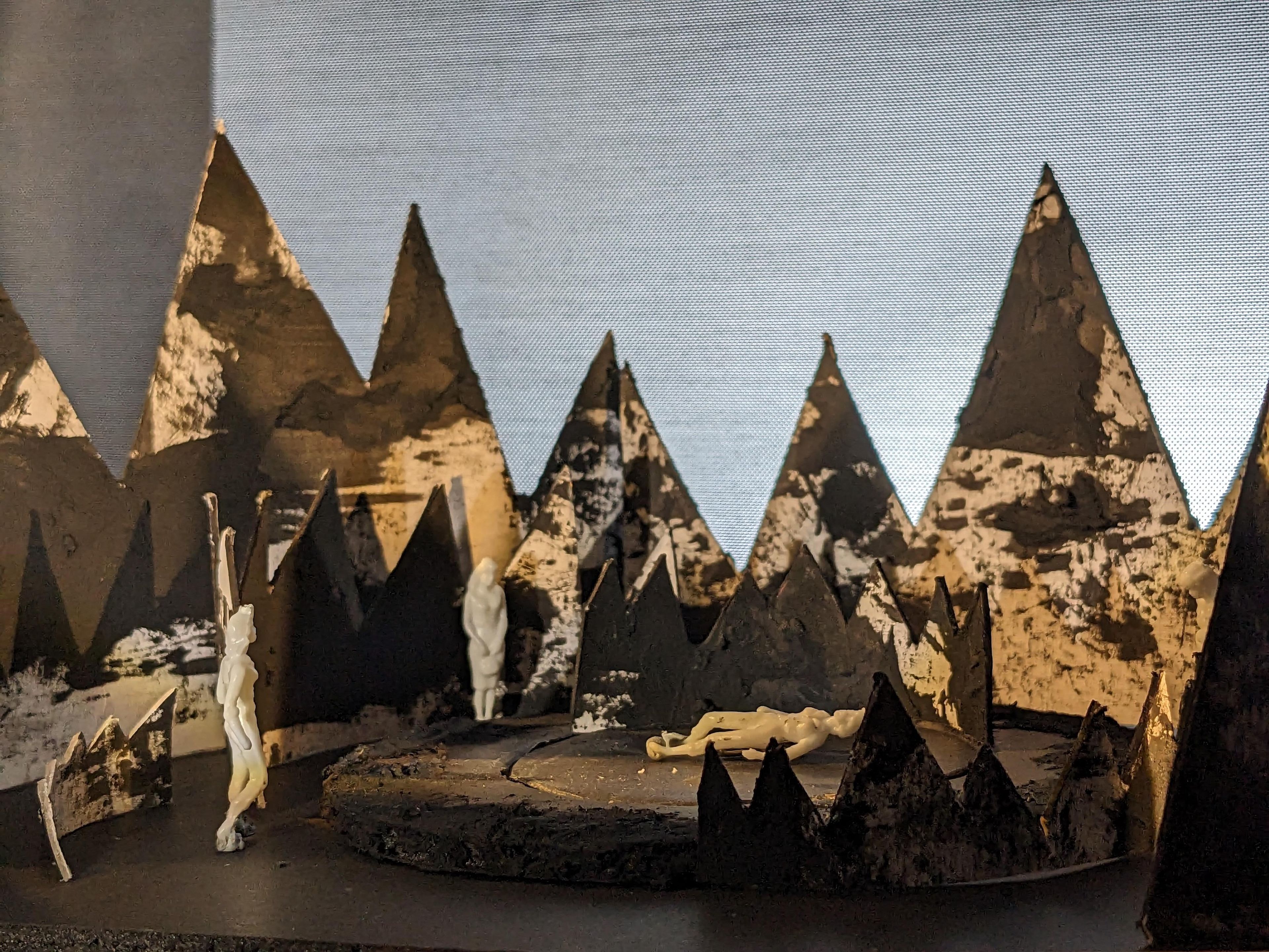 Set model with three white figures on a circular stage in front of large, black, textured mountain shapes that create a stark contrast with the lighter background.