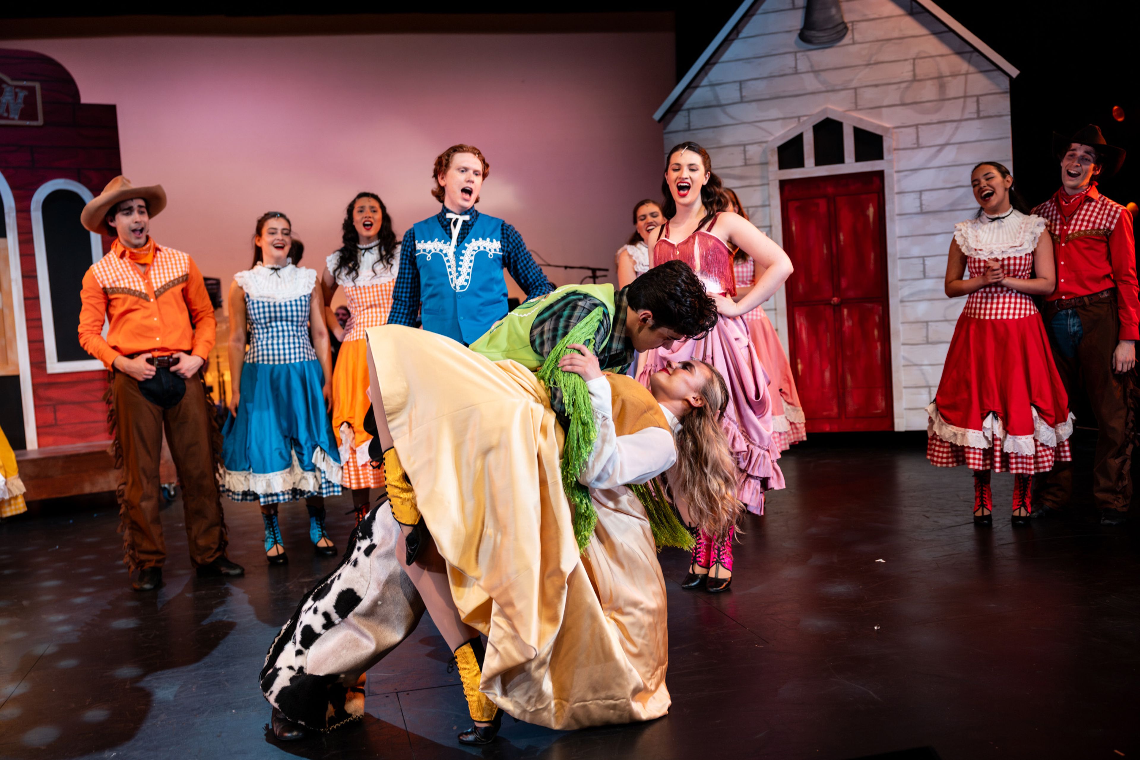 Theatre production photograph capturing a dance finale moment with performers in bright Western-inspired costumes reacting to a dramatic dip between two dancers, against a saloon and farmhouse set.