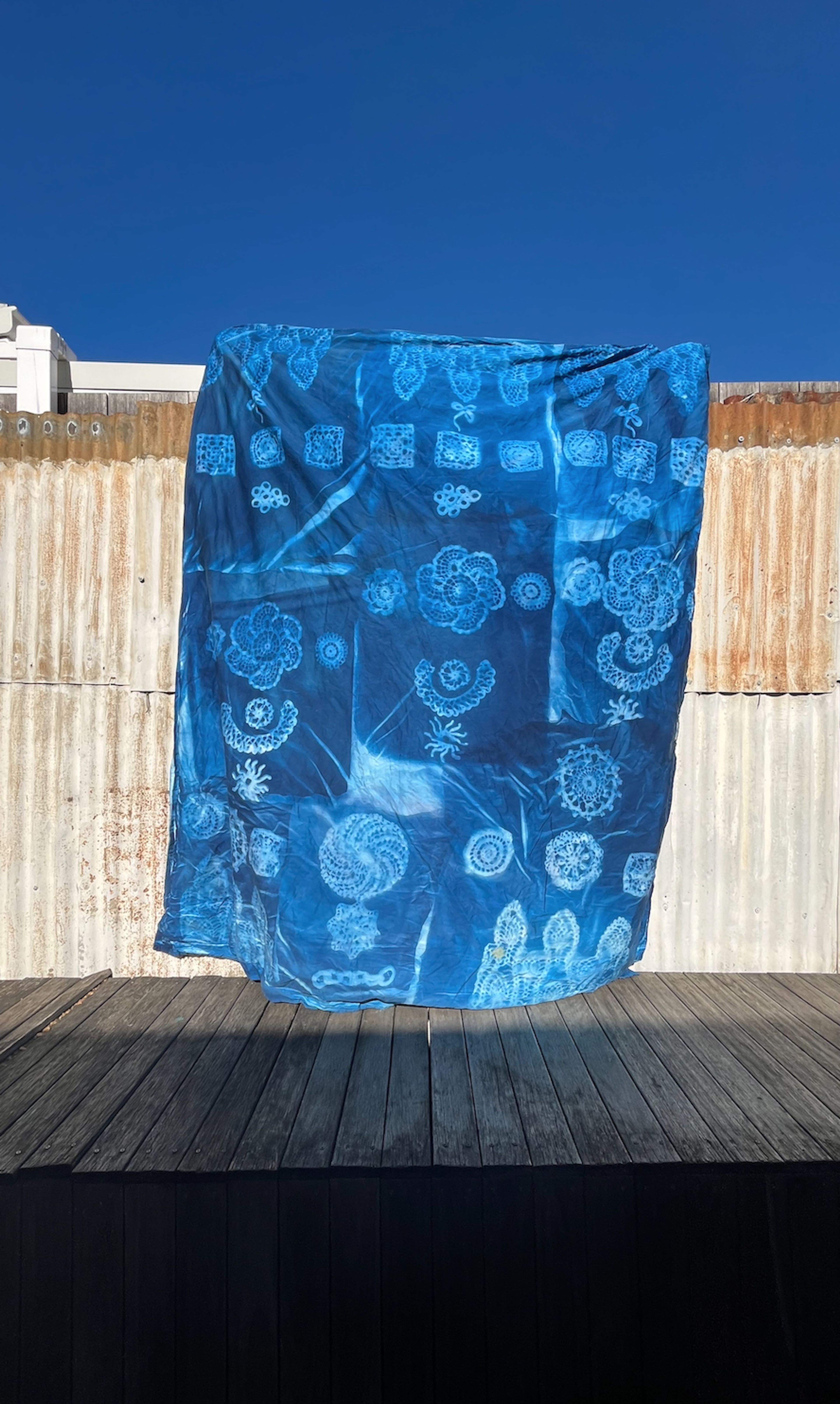 An indigo-dyed textile displayed against a blue sky and rusted corrugated metal background. The textile features intricate patterns resembling flowers, leaves, and geometric shapes, creating a natural, organic look.