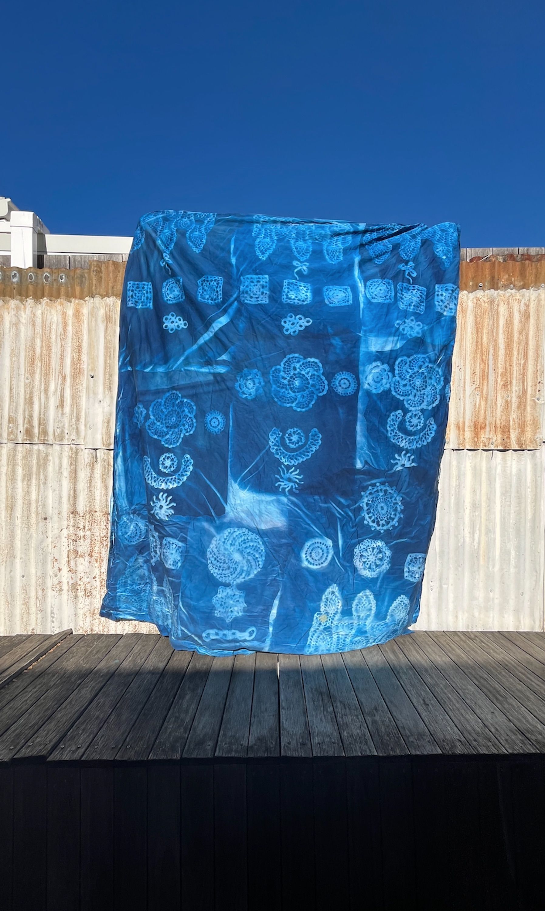 An indigo-dyed textile displayed against a blue sky and rusted corrugated metal background. The textile features intricate patterns resembling flowers, leaves, and geometric shapes, creating a natural, organic look.