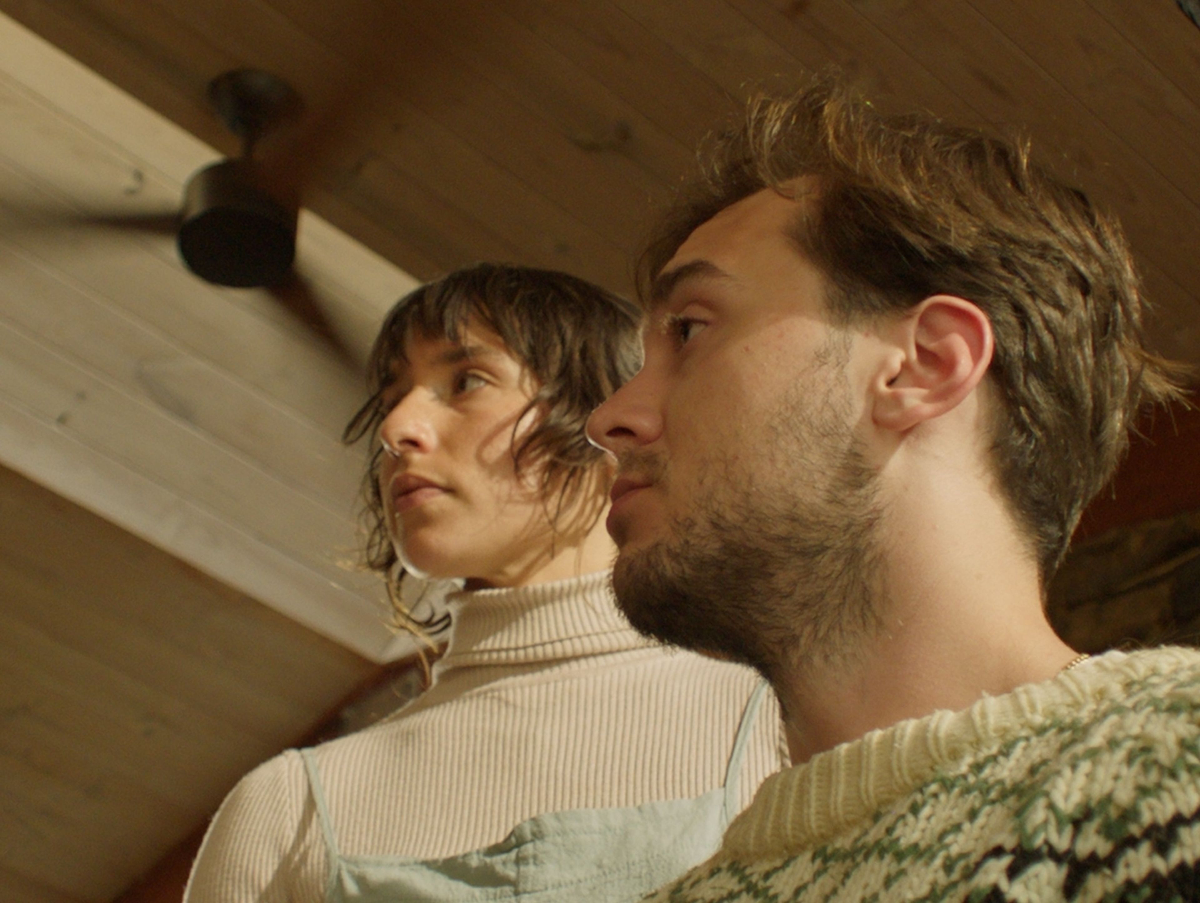 Film still featuring a close-up profile shot of two people in cream-coloured knitwear against wooden ceiling panels, shot in warm interior lighting.