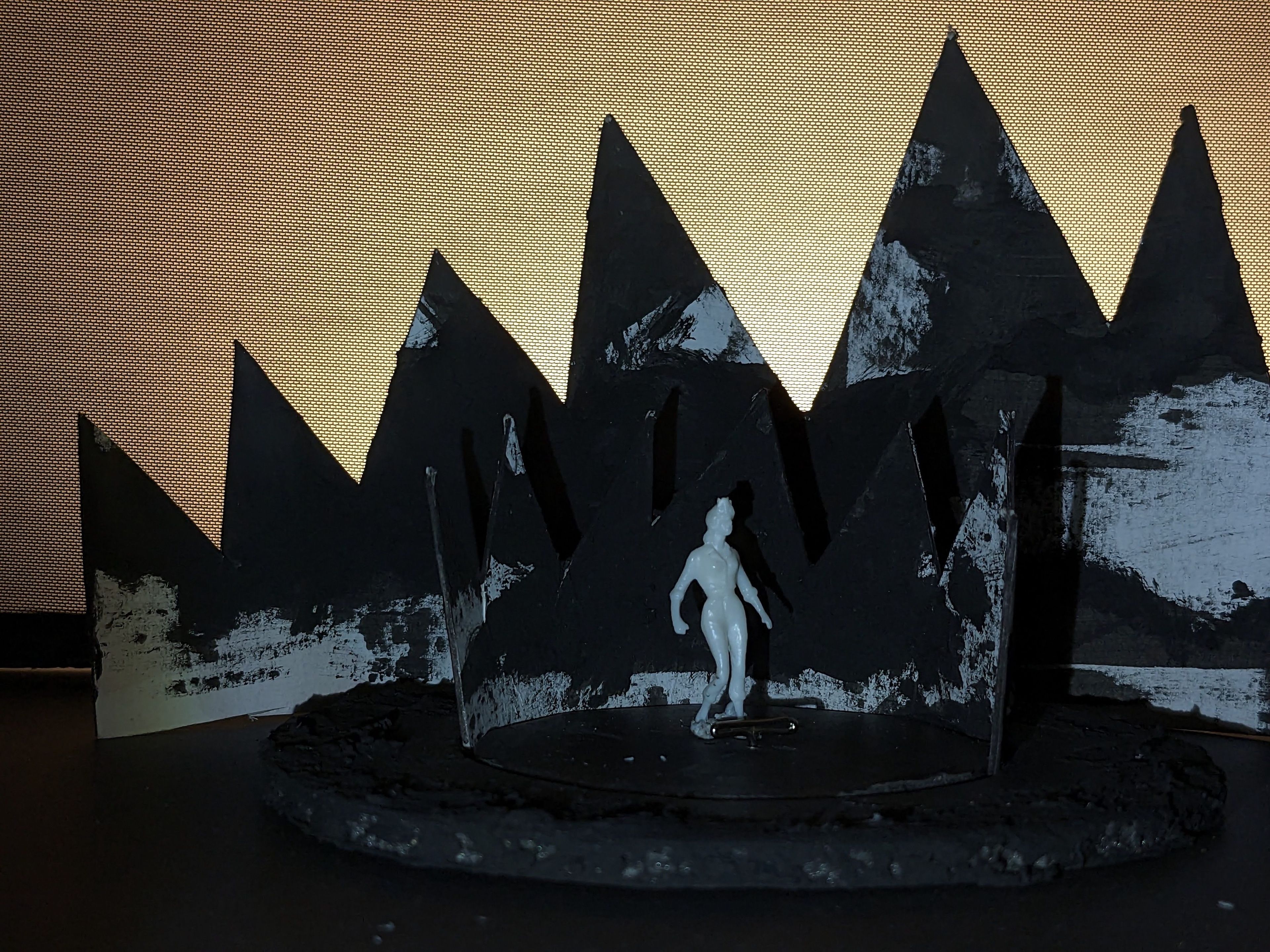 Scale model depicting a white figure standing on a round, textured stage surrounded by black, pointed mountain shapes against a warm, gradient background.