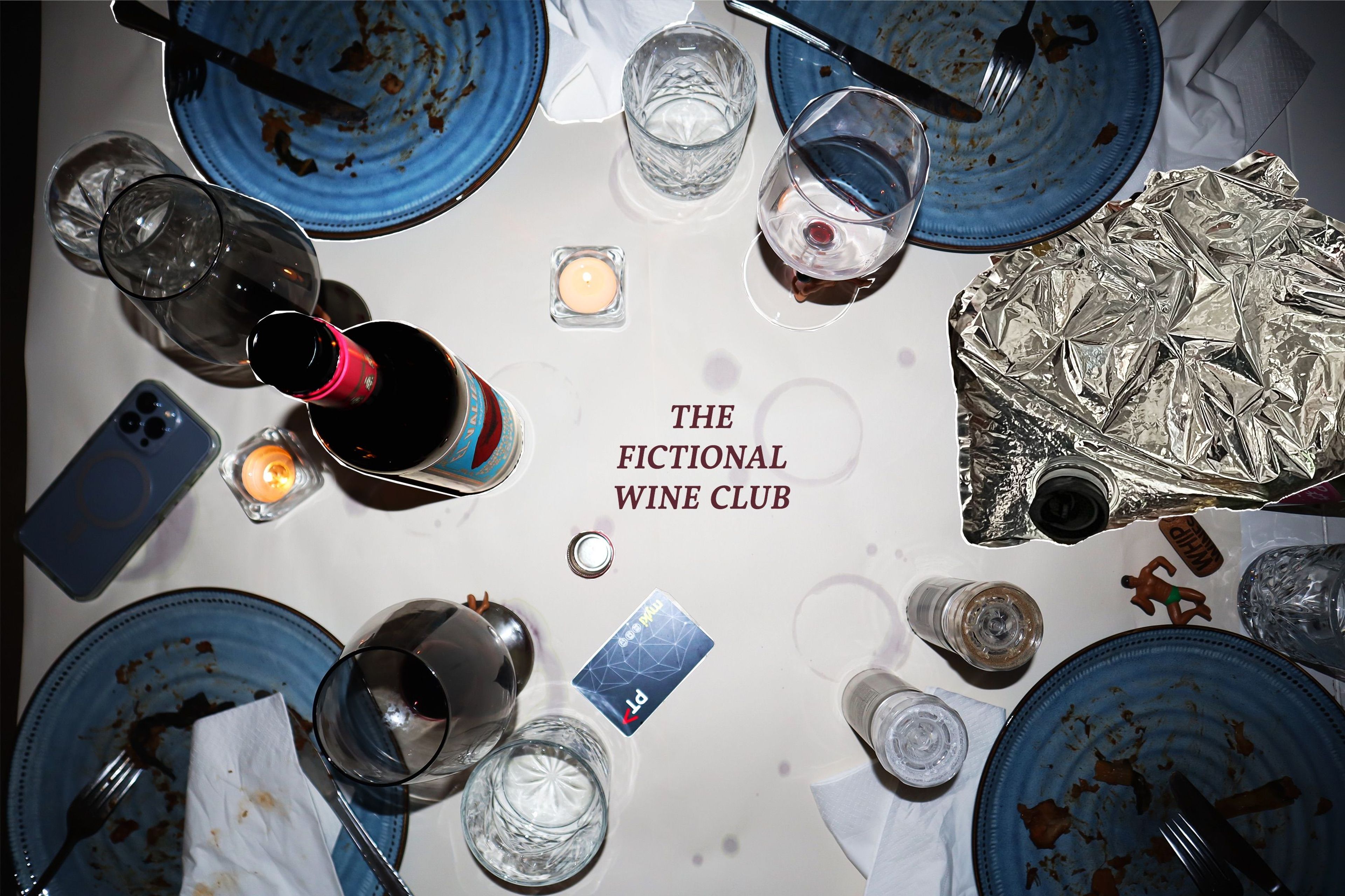 Overhead view of a dinner table with remnants of food on blue plates, half-empty wine glasses, candles, and an empty wine bottle, with the text "The Fictional Wine Club" printed on the tablecloth.