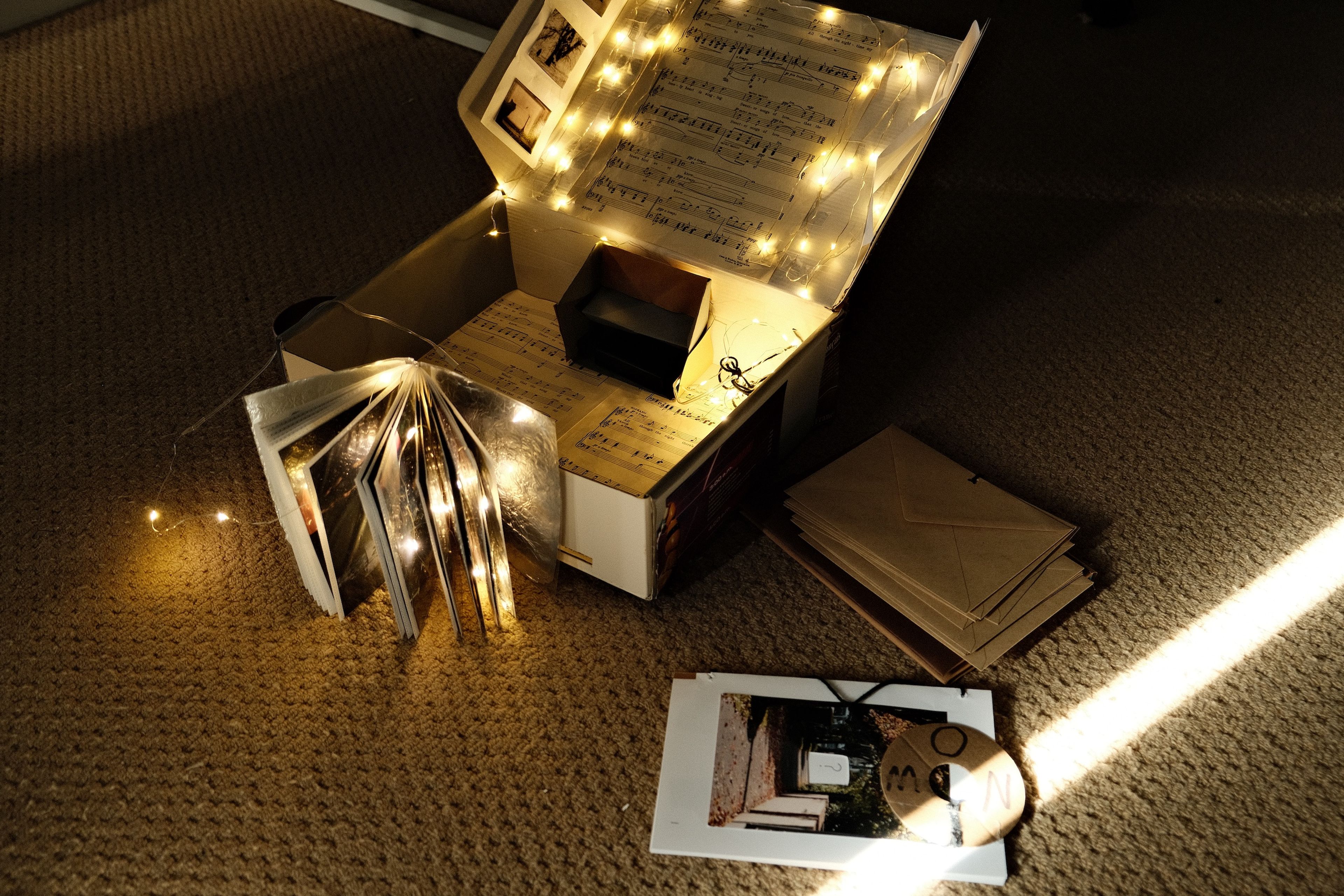 Open white box displaying sheet music interior, illuminated by fairy lights. Features a creatively lit photo book with pages fanned out and lights woven through, creating an intimate atmospheric composition on beige carpet.