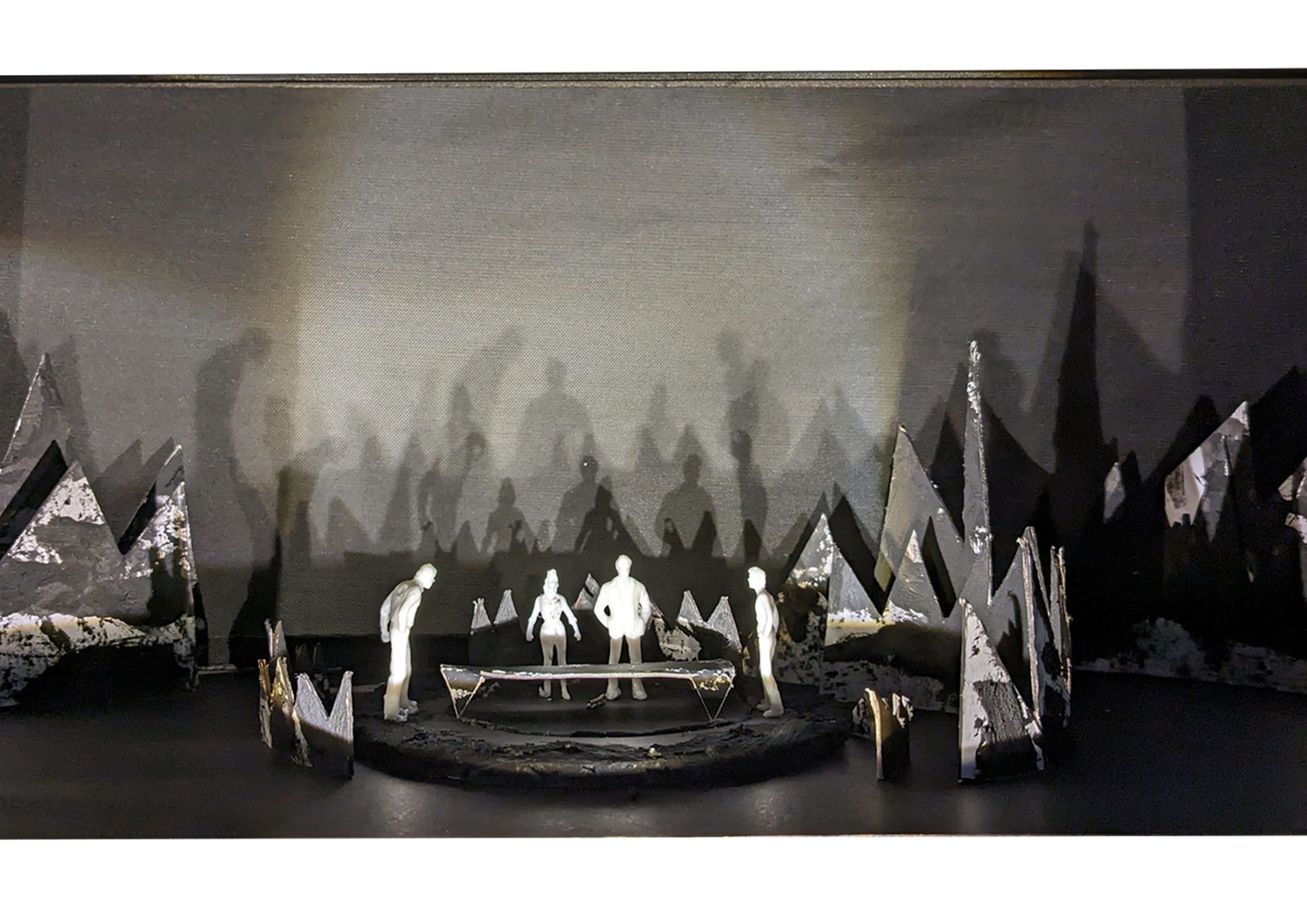 Scale model of a stage with four white figures surrounding a central platform, creating shadow effects against a backdrop of black mountains.