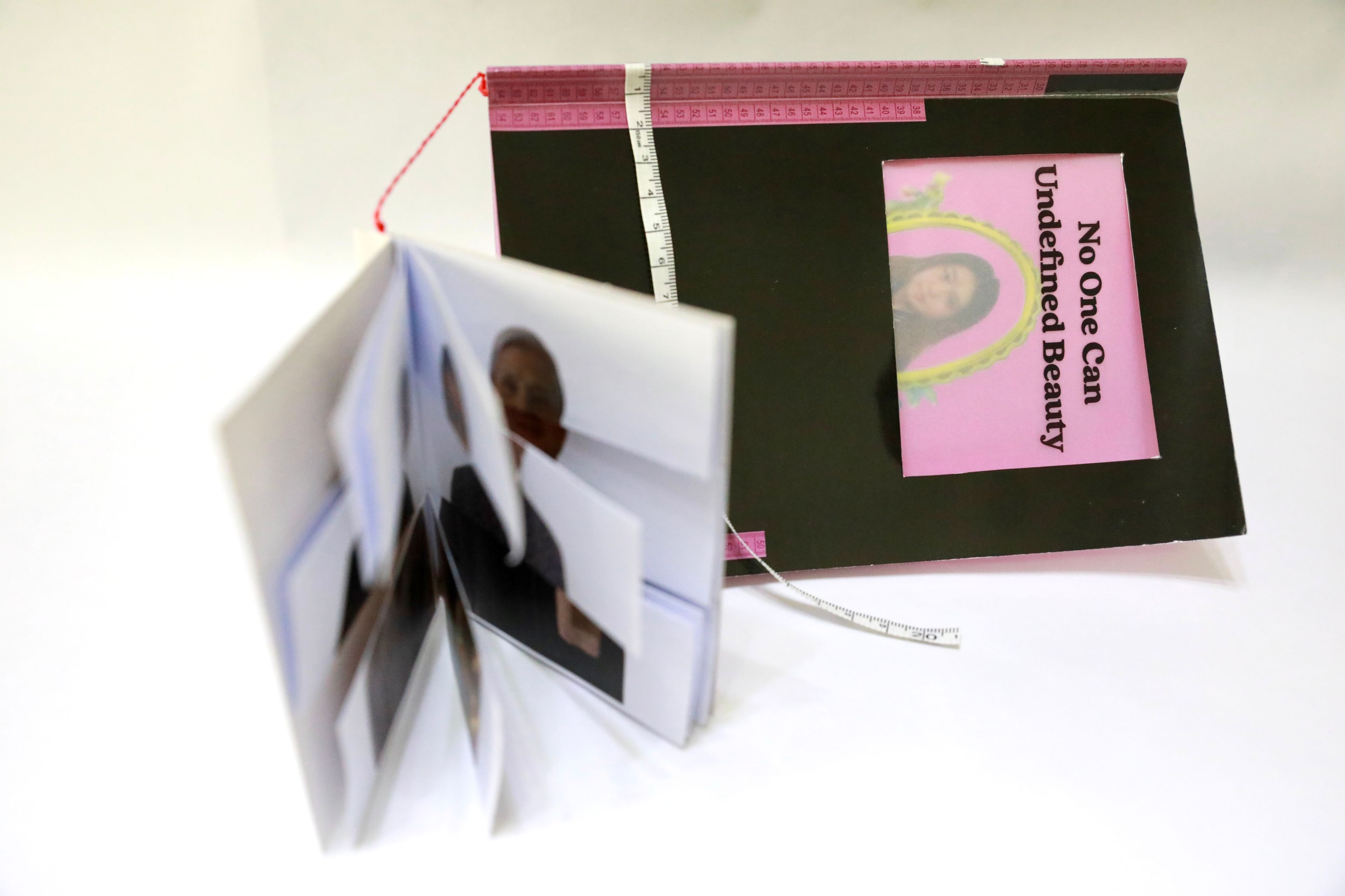 Handmade art book with a black cover, featuring a pink-framed portrait on the right page with the text 'No One Can Undefined Beauty.' A pink measuring tape decorates the edges, with pages of overlapping, blurred portraits extending outward.