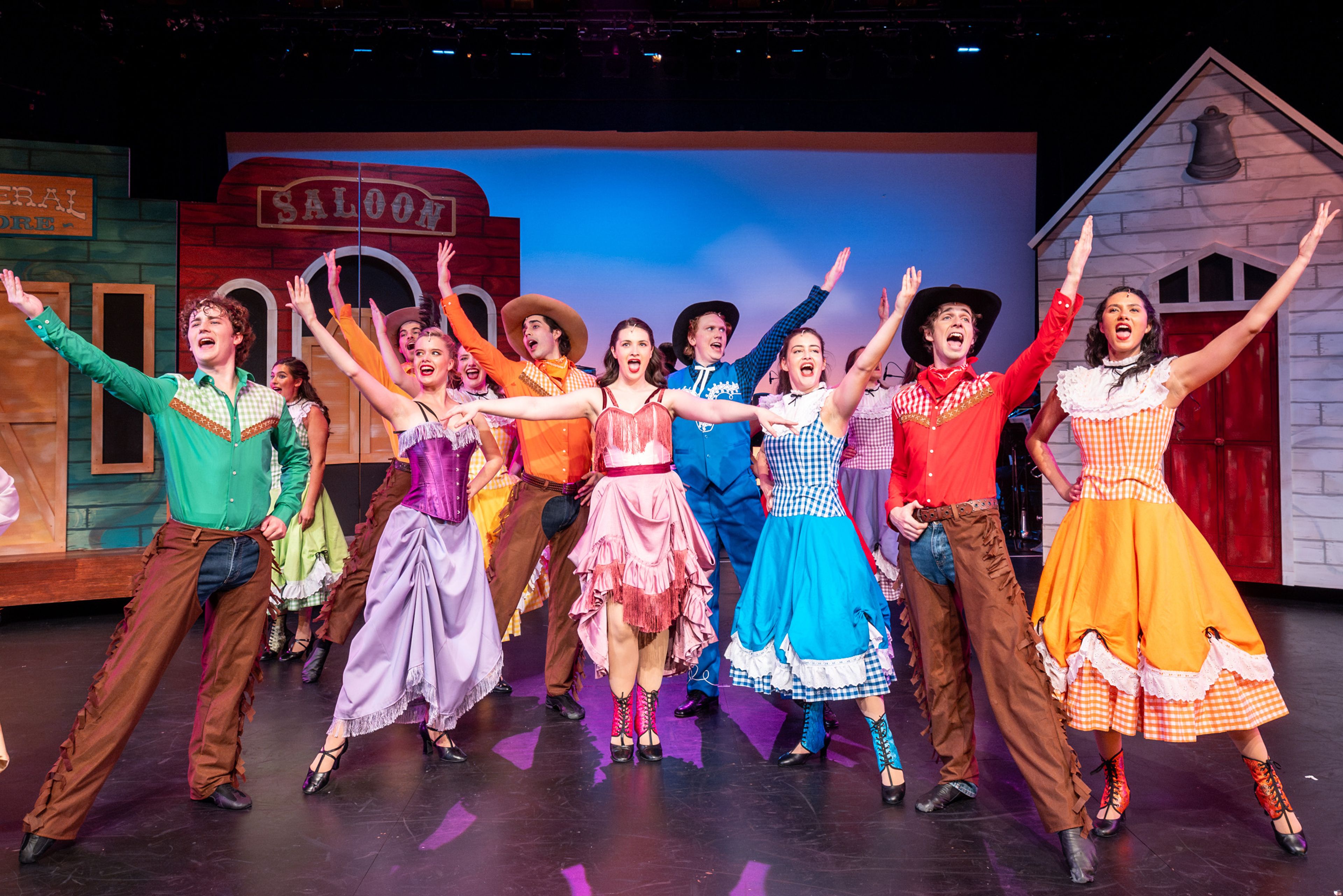 Theatre production photograph showing performers in bright 1950s outfits, with women in full skirts and men in Western wear, joyfully singing in front of a period-style saloon set.