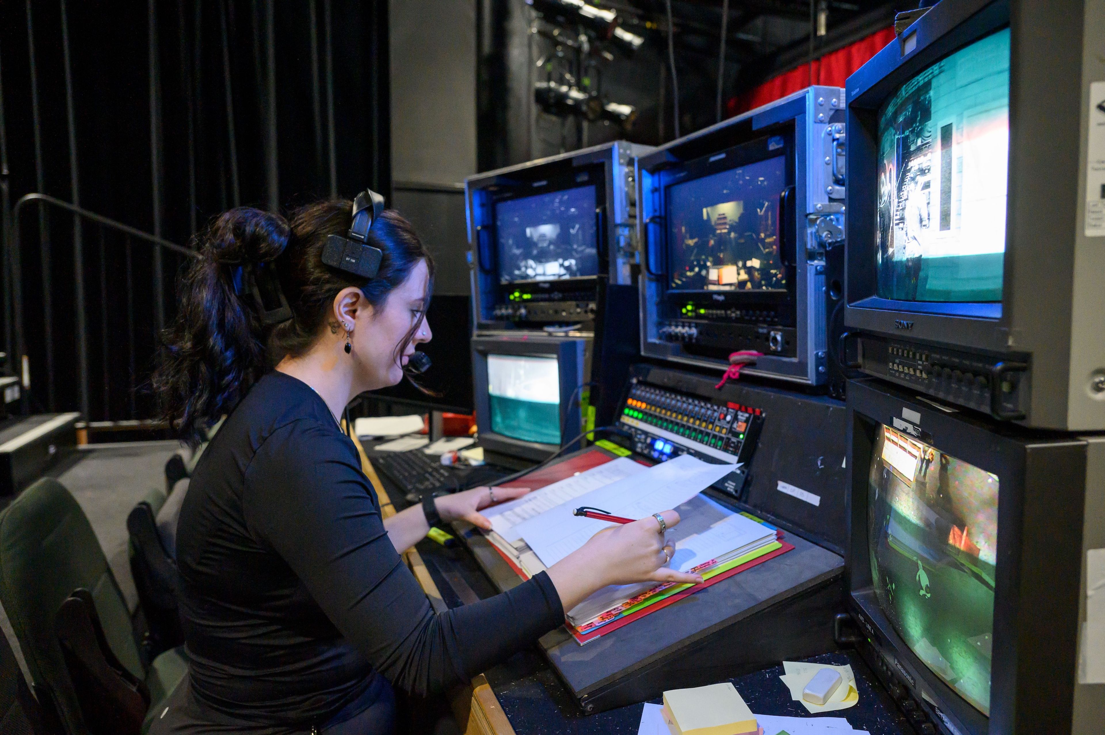 Close-up of a crew member in a headset focused on their script at a backstage console with multiple monitors showing live stage views.