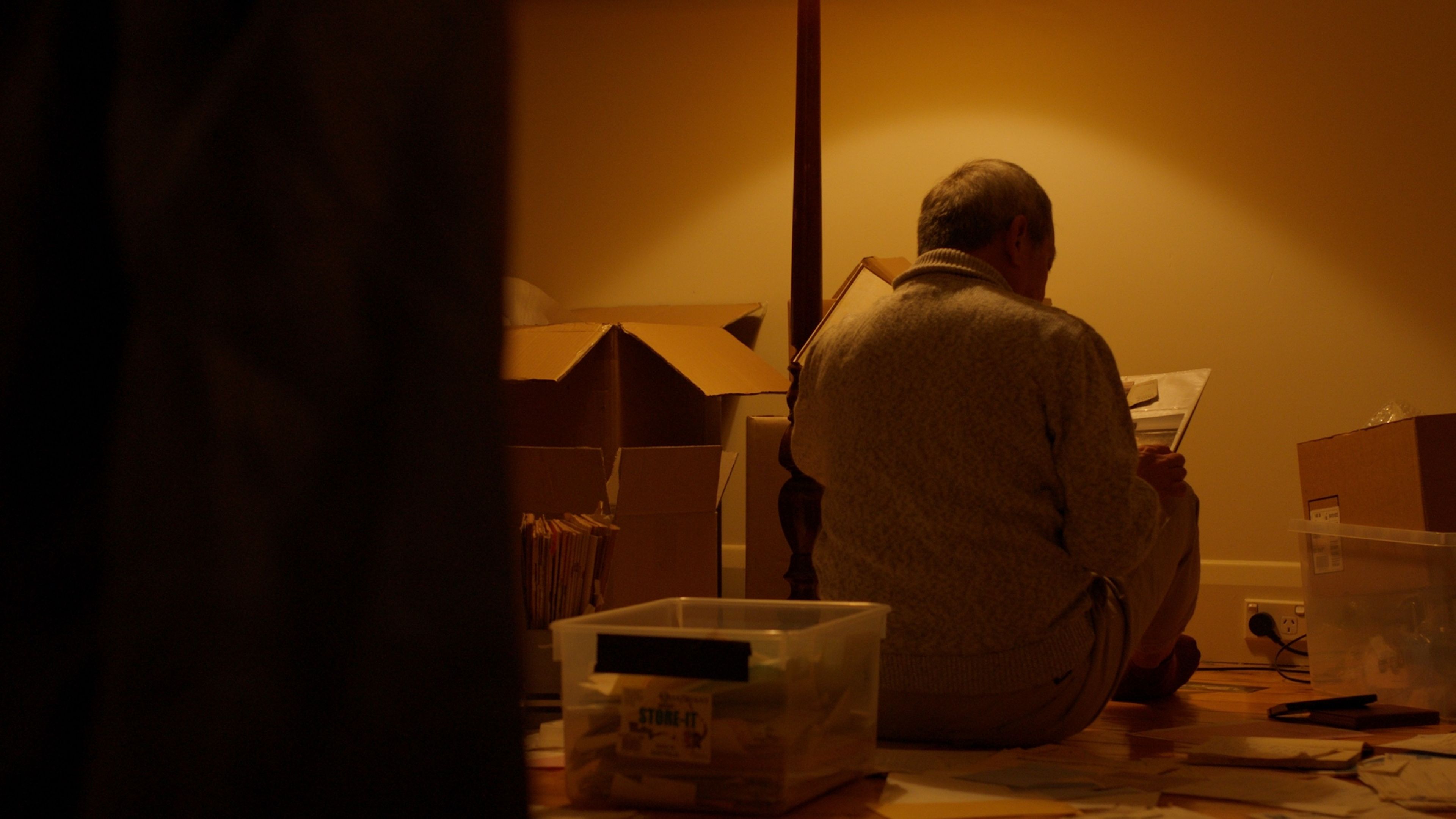 Film still showing a man in a beige jumper sitting amongst cardboard boxes and storage containers, bathed in warm amber lighting