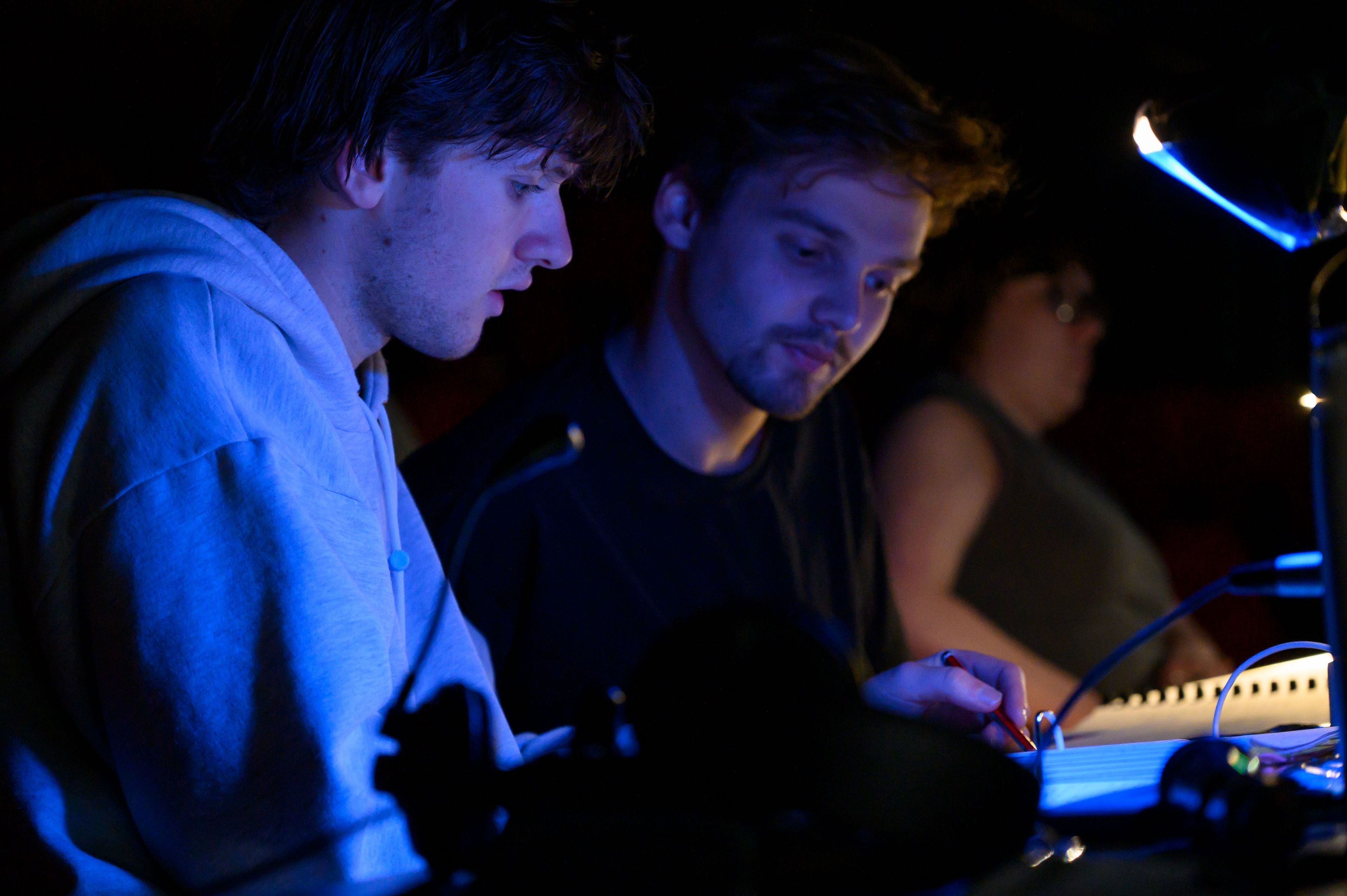 Two crew members work together, intently studying a script or control panel under dim blue lighting.