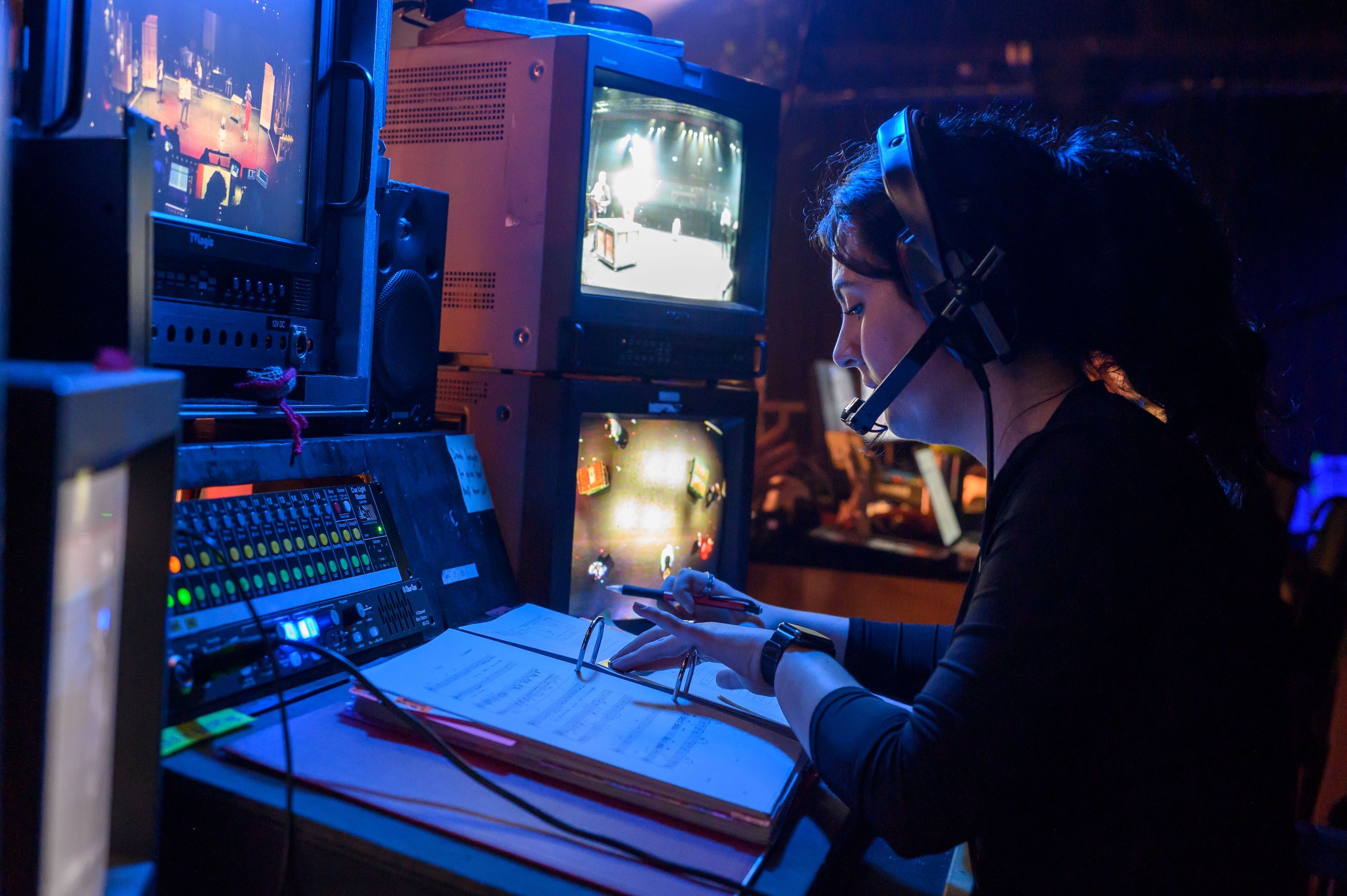 Close-up of a stage manager in a headset, monitoring multiple screens displaying live footage from the stage. She is focused on her notes and equipment.