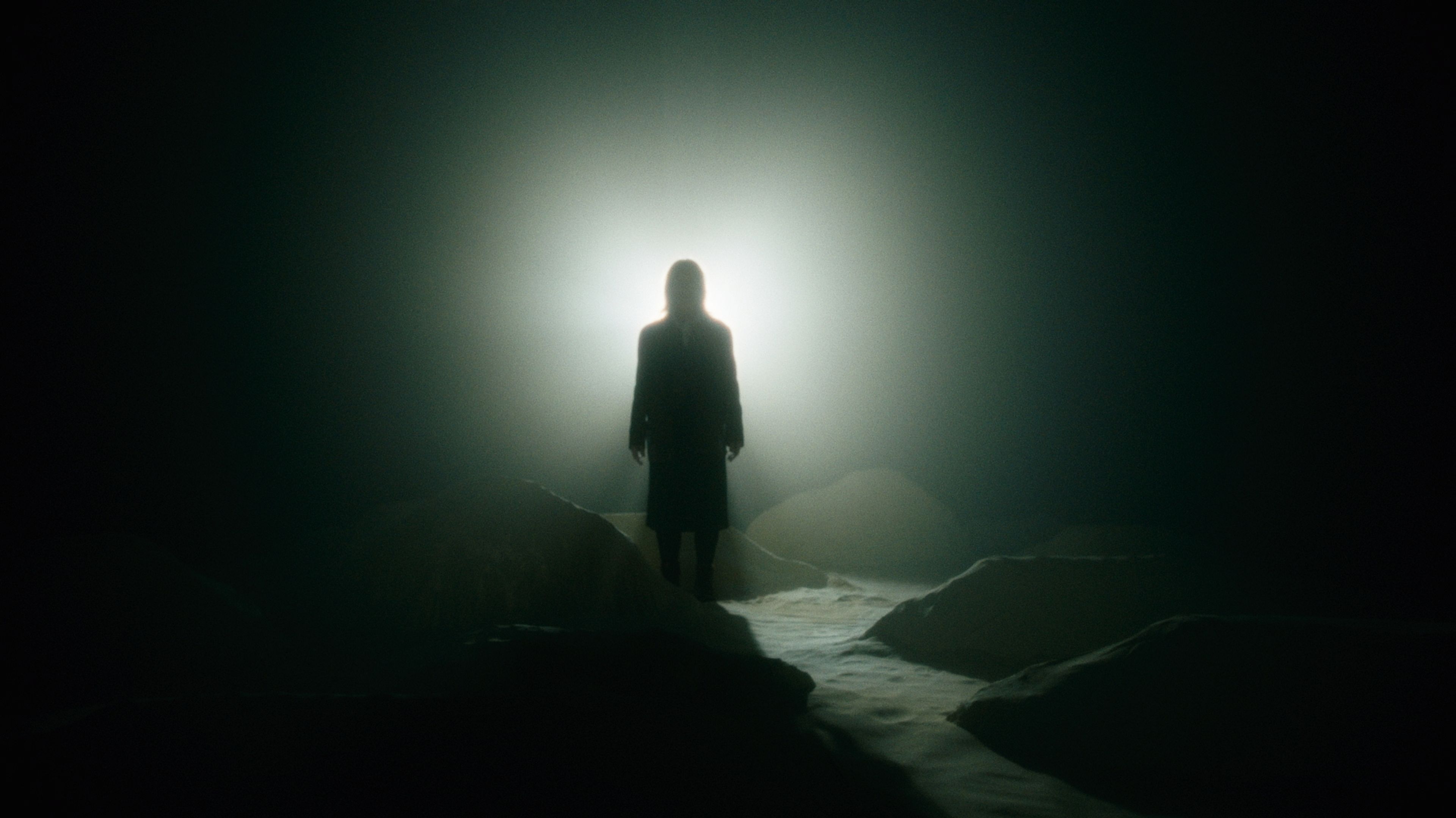 Film still showing a silhouetted figure standing on a rocky outcrop against a glowing white light, surrounded by mist and shadows.