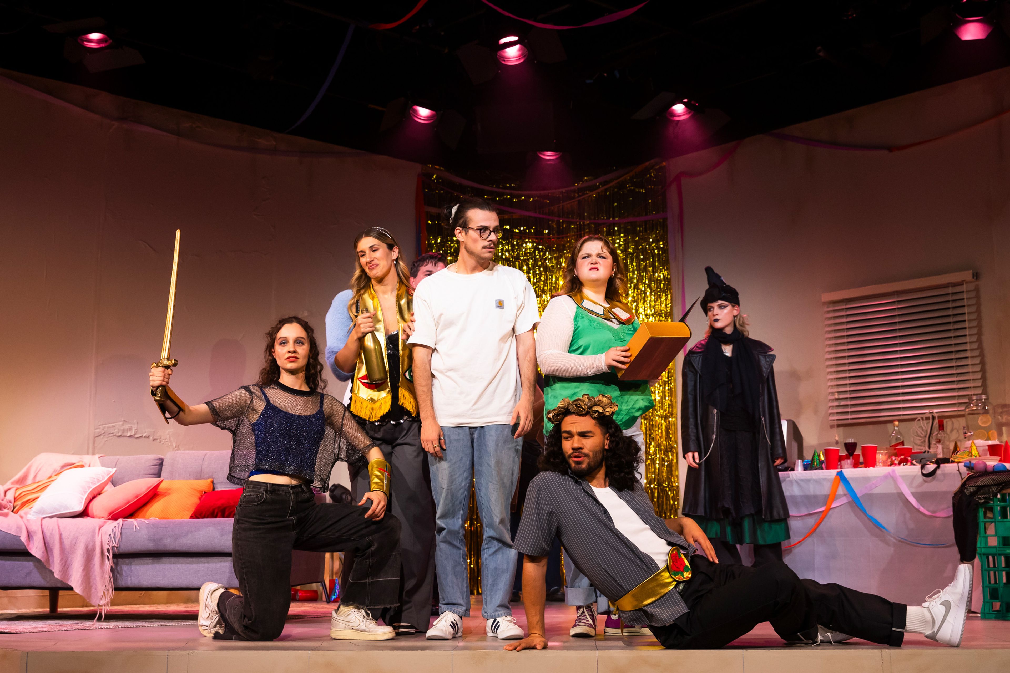 Theatre production photograph capturing a theatrical moment with a group of performers in party costumes including one wielding a prop sword, against a gold tinsel backdrop.