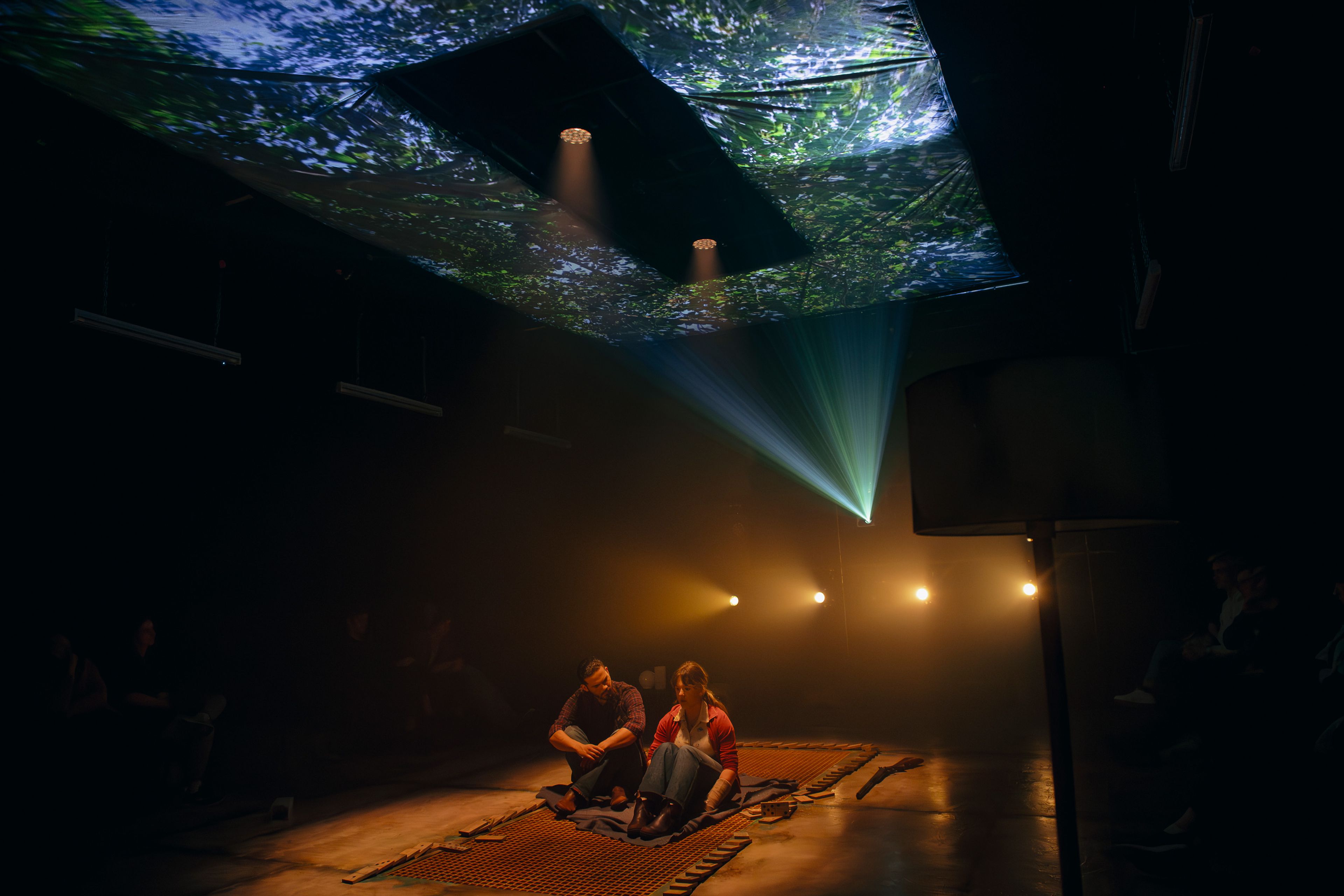 Theatre production photograph showing two performers sitting on an orange mat under atmospheric lighting, with tree projections visible on the ceiling and amber footlights creating depth.