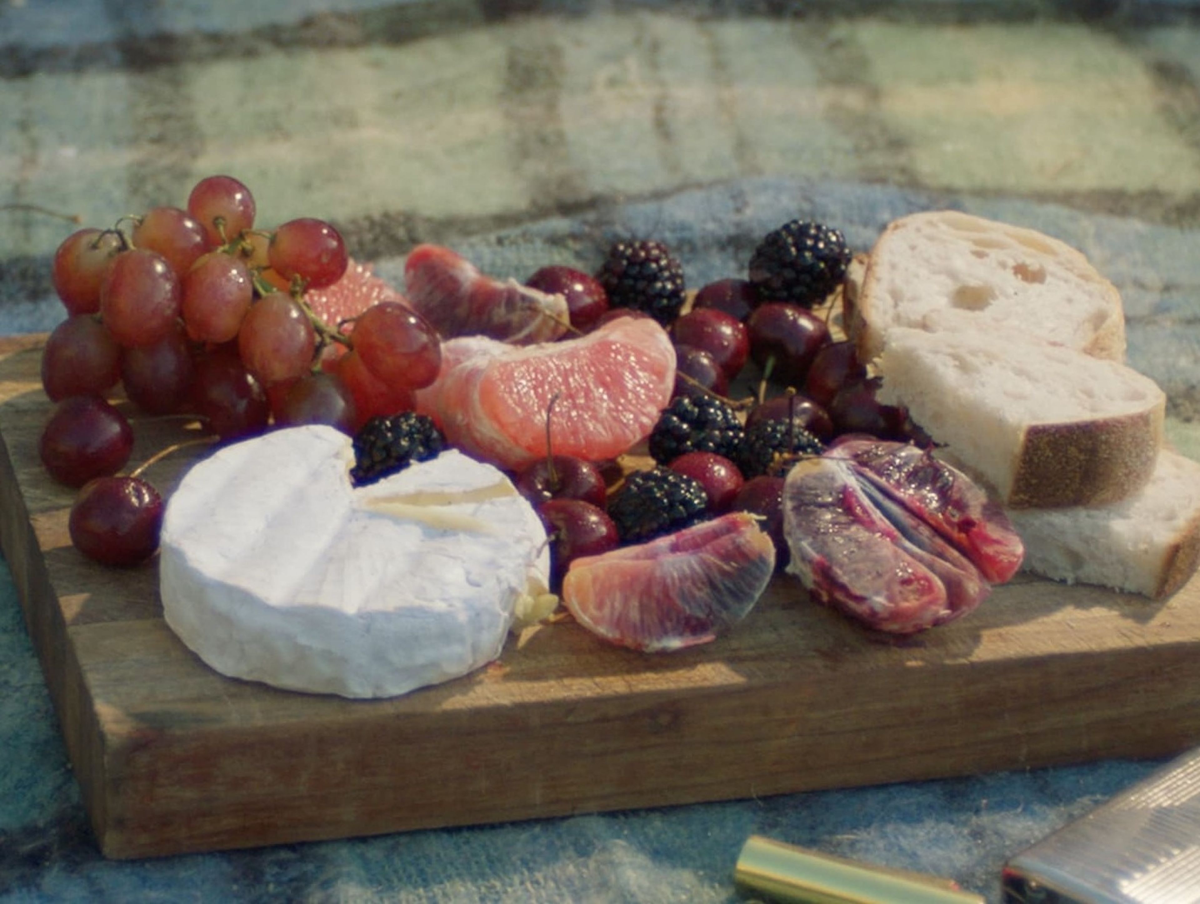 Film still of a rustic wooden cheese board featuring brie, grapes, blood oranges, blackberries and crusty bread, arranged artfully and shot in natural light.