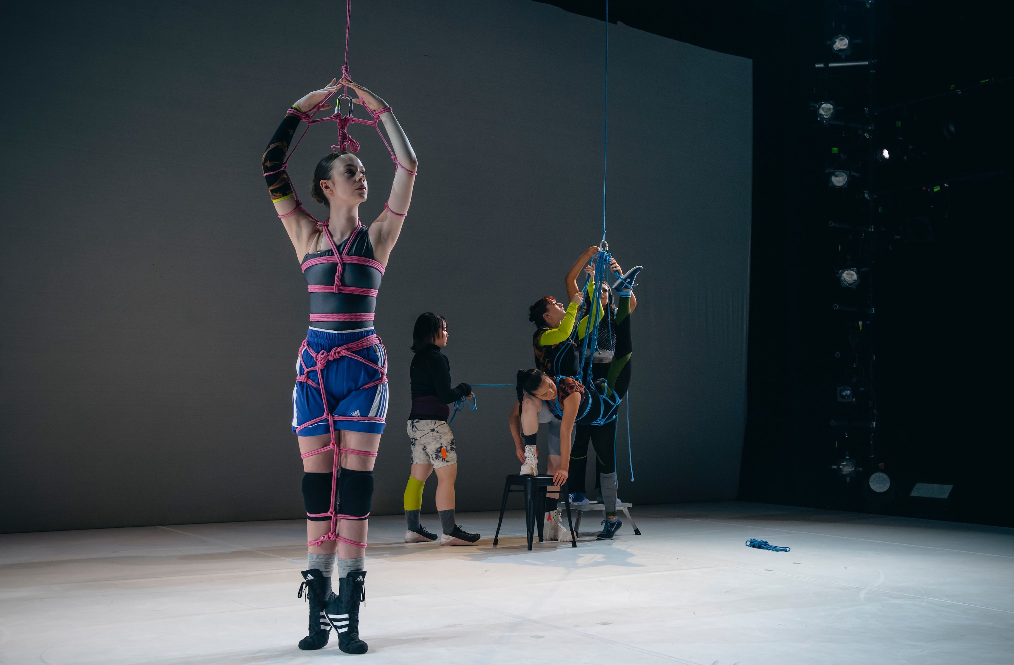 A dancer in blue shorts and black sports top bound in pink ropes in a standing pose, while others work with ropes and chairs in the background.