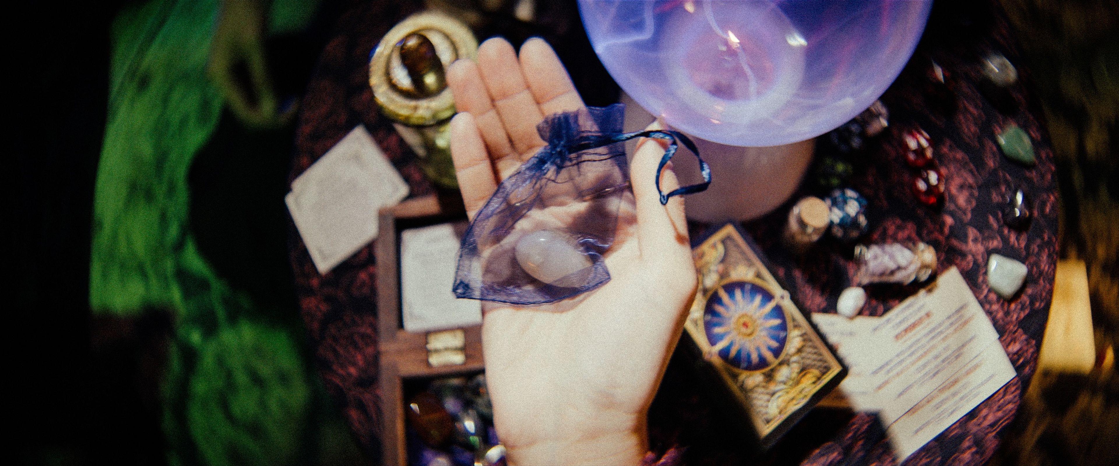 Close-up film still of mystical props including tarot cards, crystal balls, and organza pouches arranged on a patterned tablecloth, lit dramatically with coloured gels