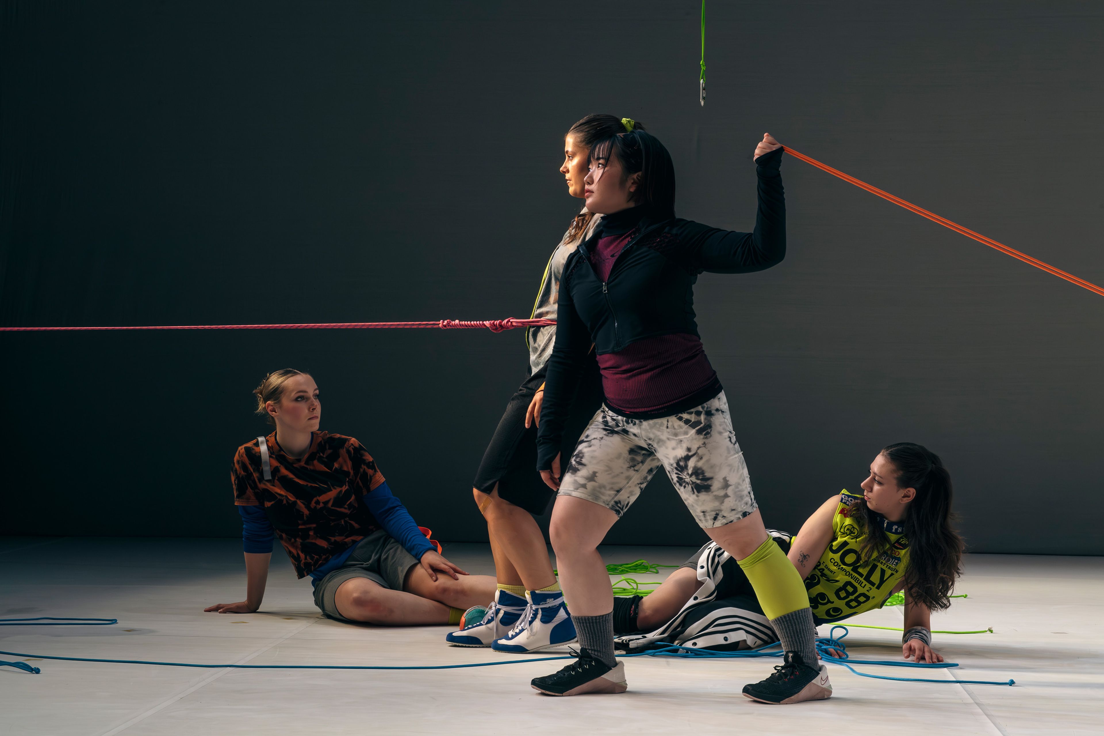 Dance production photograph with performers arranged in various poses, manipulating coloured ropes to form interconnected lines across the stage.