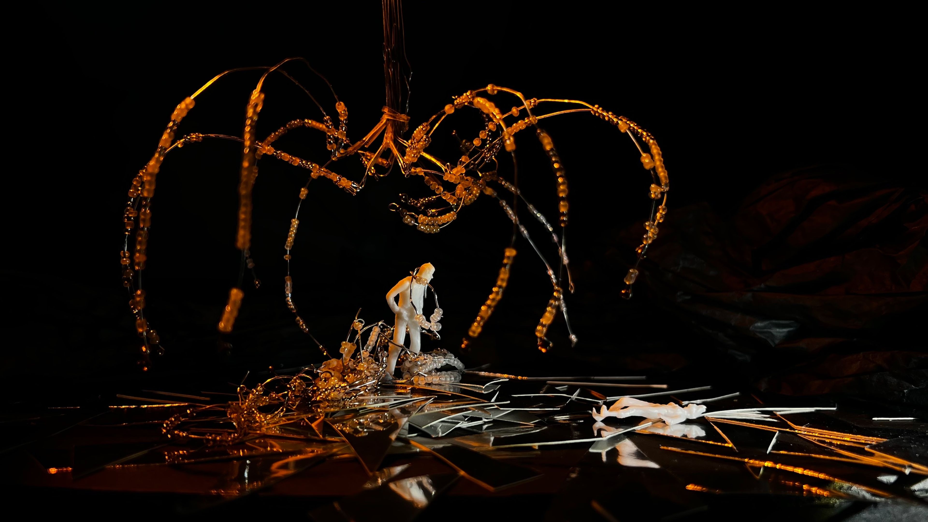 Scale model featuring a central white figure standing on shattered mirror-like surfaces, beneath an arching amber-coloured beaded structure, dramatically lit from above