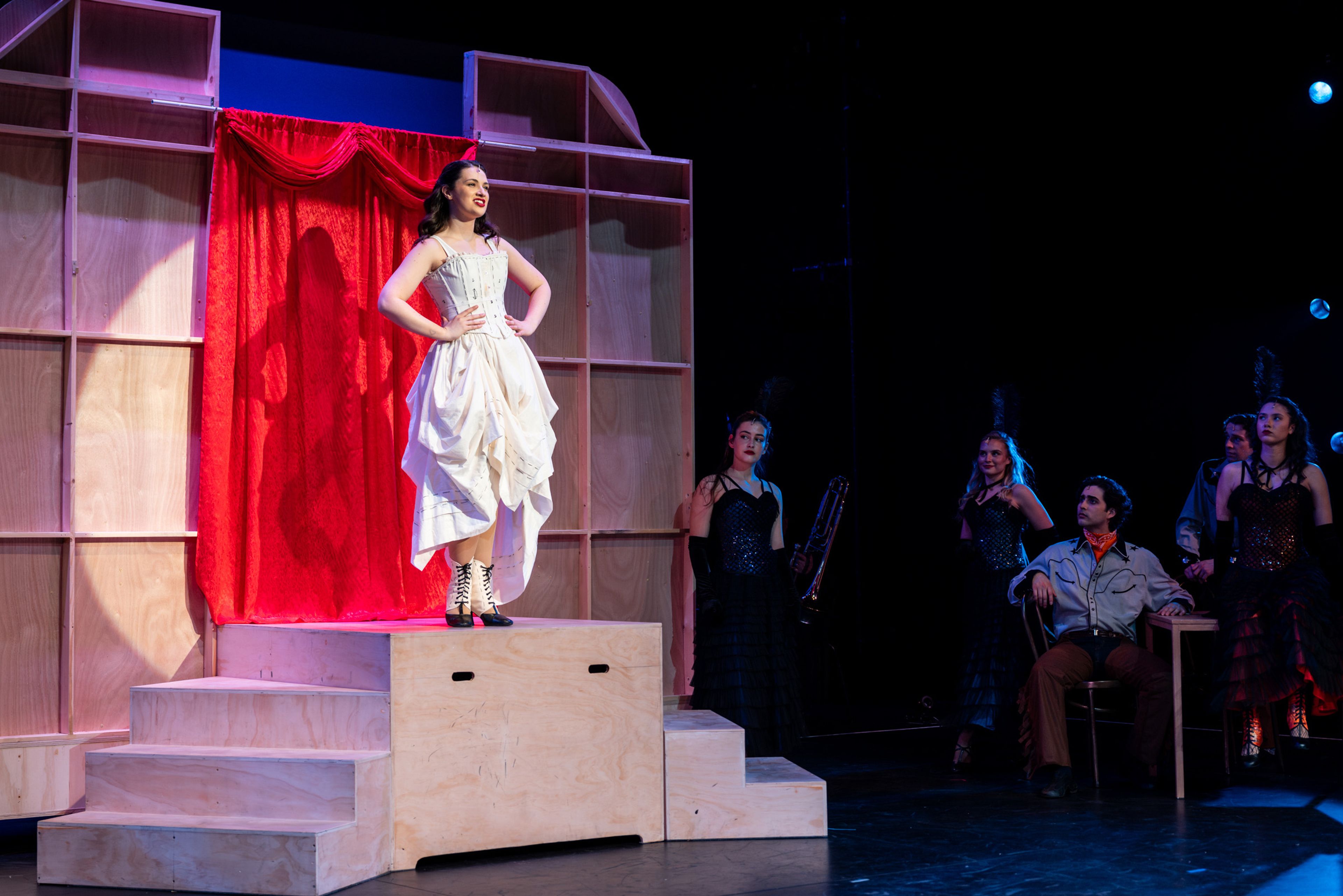 Theatre production photograph showing a performer in a white corset dress on stepped wooden platforms, with a red curtain backdrop and other cast members partially visible in darker lighting.