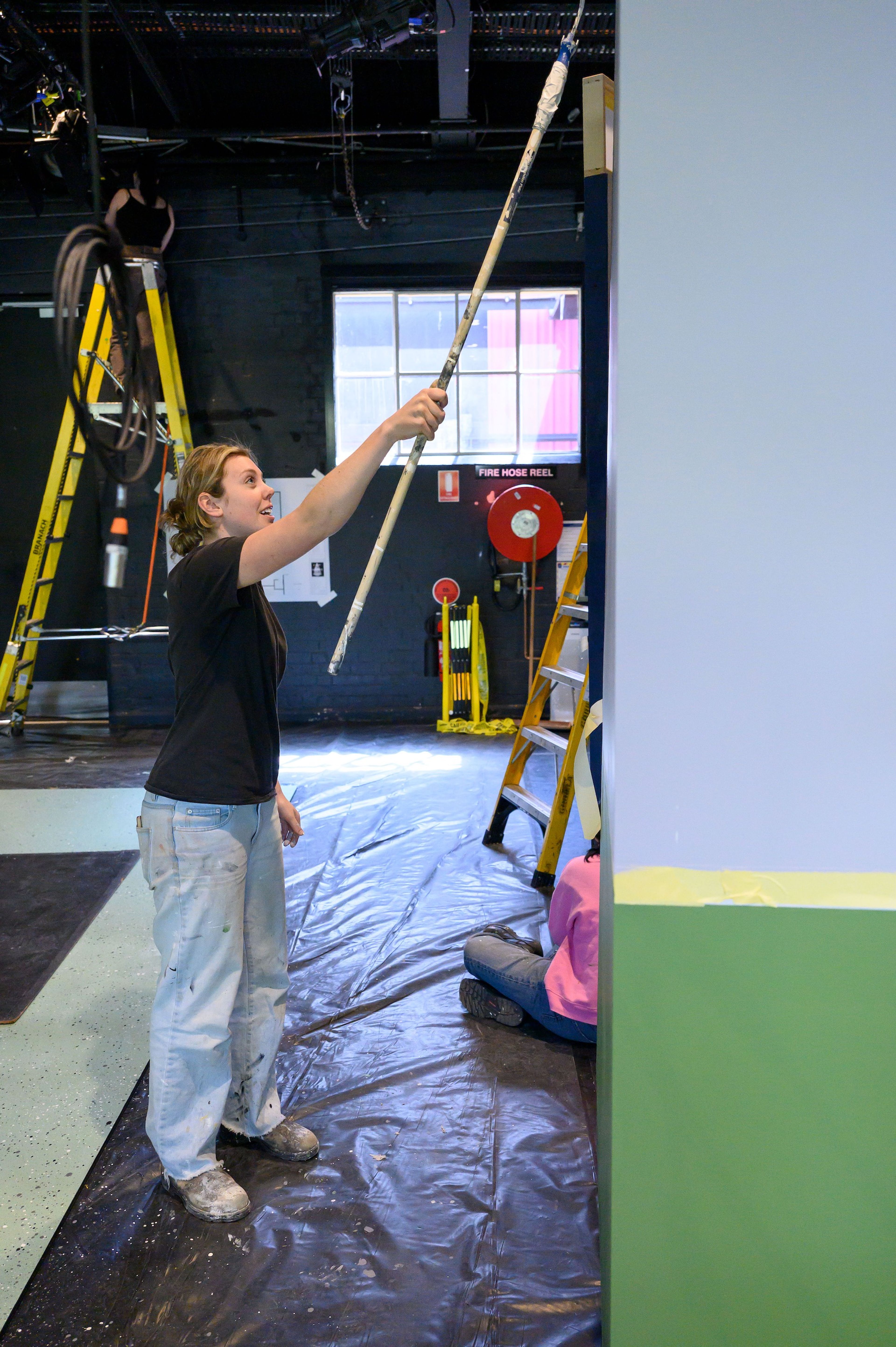 Behind-the-scenes photograph of a person painting a tall set piece with a roller, with stage equipment and a ladder visible in the background.