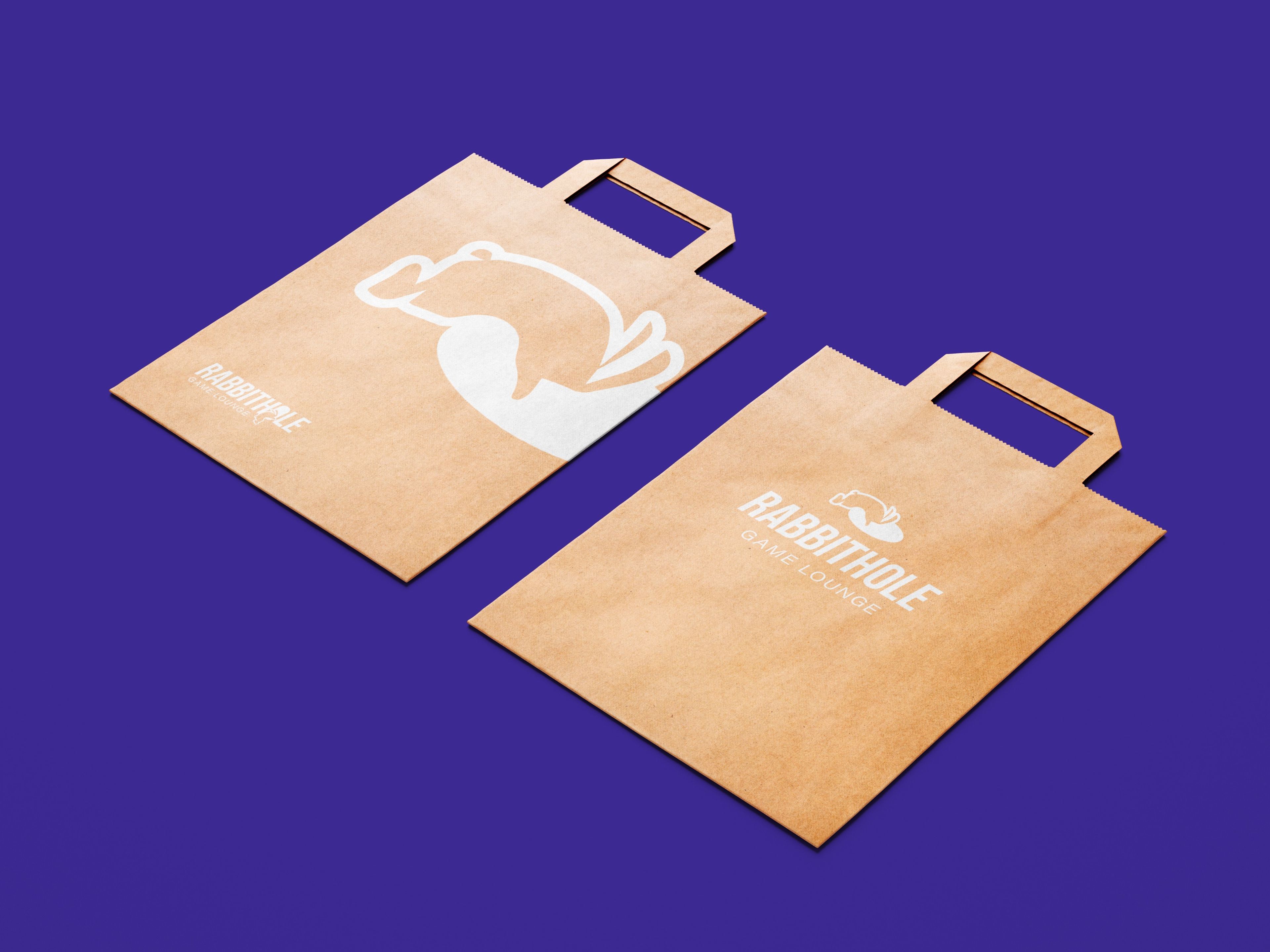 Two brown paper bags with cut-out handles, designed for 'Rabbithole Game Lounge.' The bags feature the brand’s logo of a stylised rabbit alongside the lounge’s name in white, set against a rich purple background.