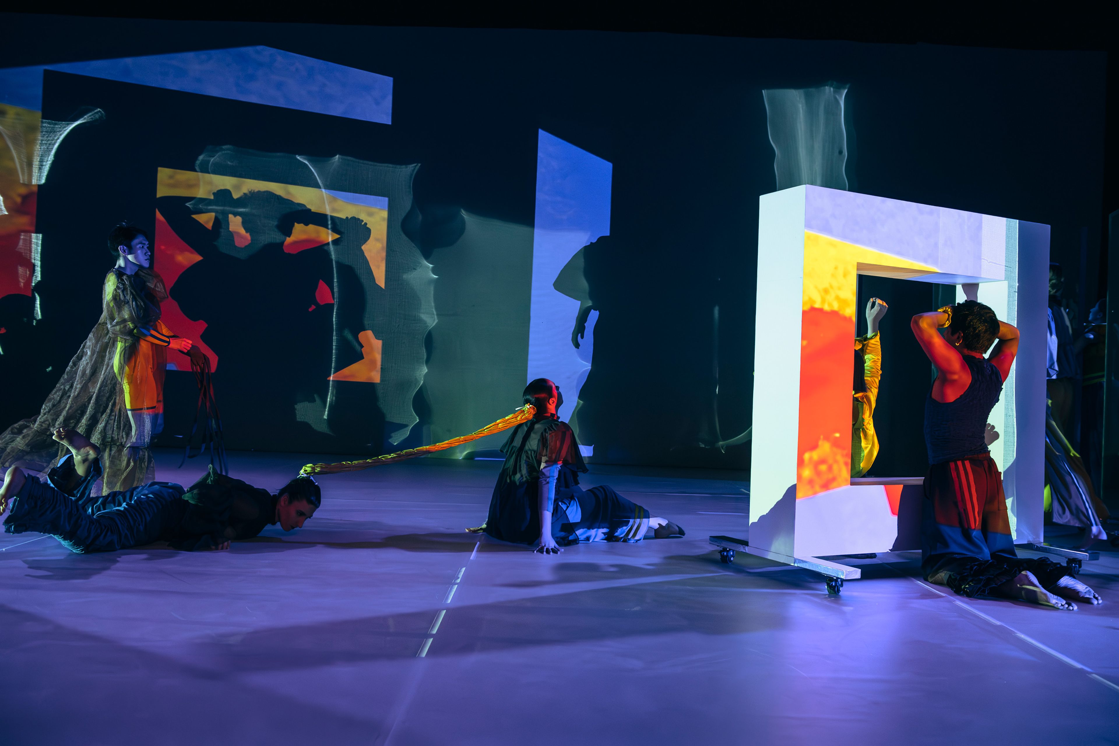 Dance production photograph of dancers on the floor and interacting with panels, illuminated by vibrant, projected colours.